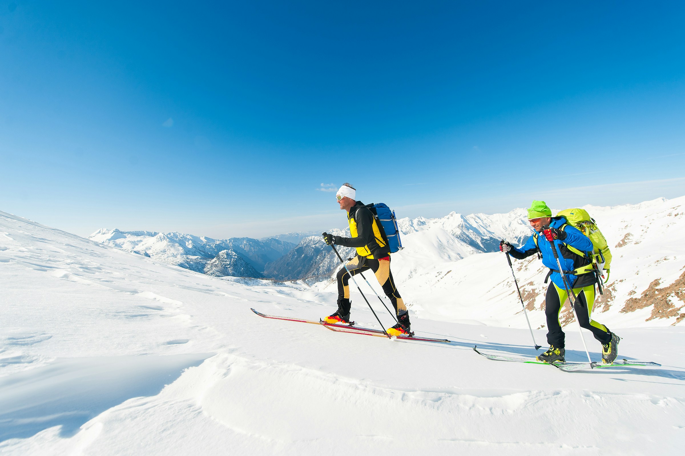 Two people on skis walk across snow-covered mountains during a ski trip with a clear blue sky in the background