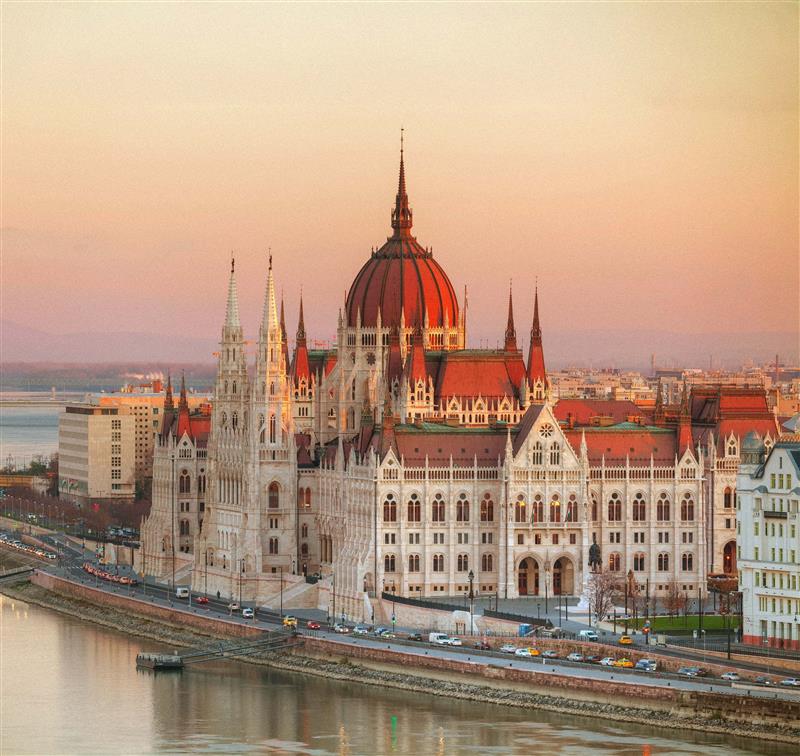 Hungarian Parliament Building at sunset, overlooking the Danube River in Budapest, with warm pastel sky tones and surrounding cityscape.