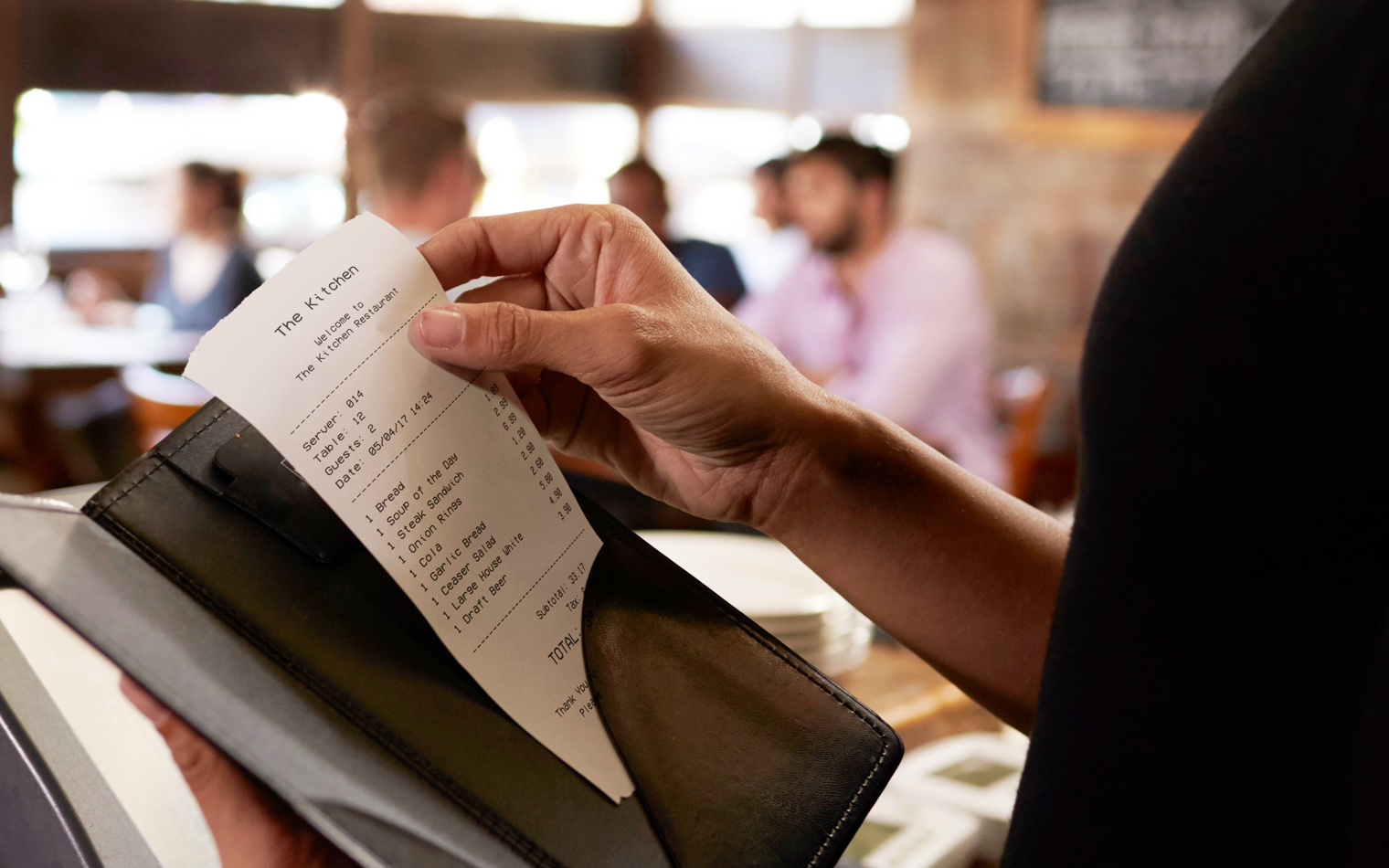 Person håller en restaurangnota i en läderhållare medan de sitter vid ett bord i en restaurang. Flera människor syns i bakgrunden, samt ett oskarpt restauranginteriör.