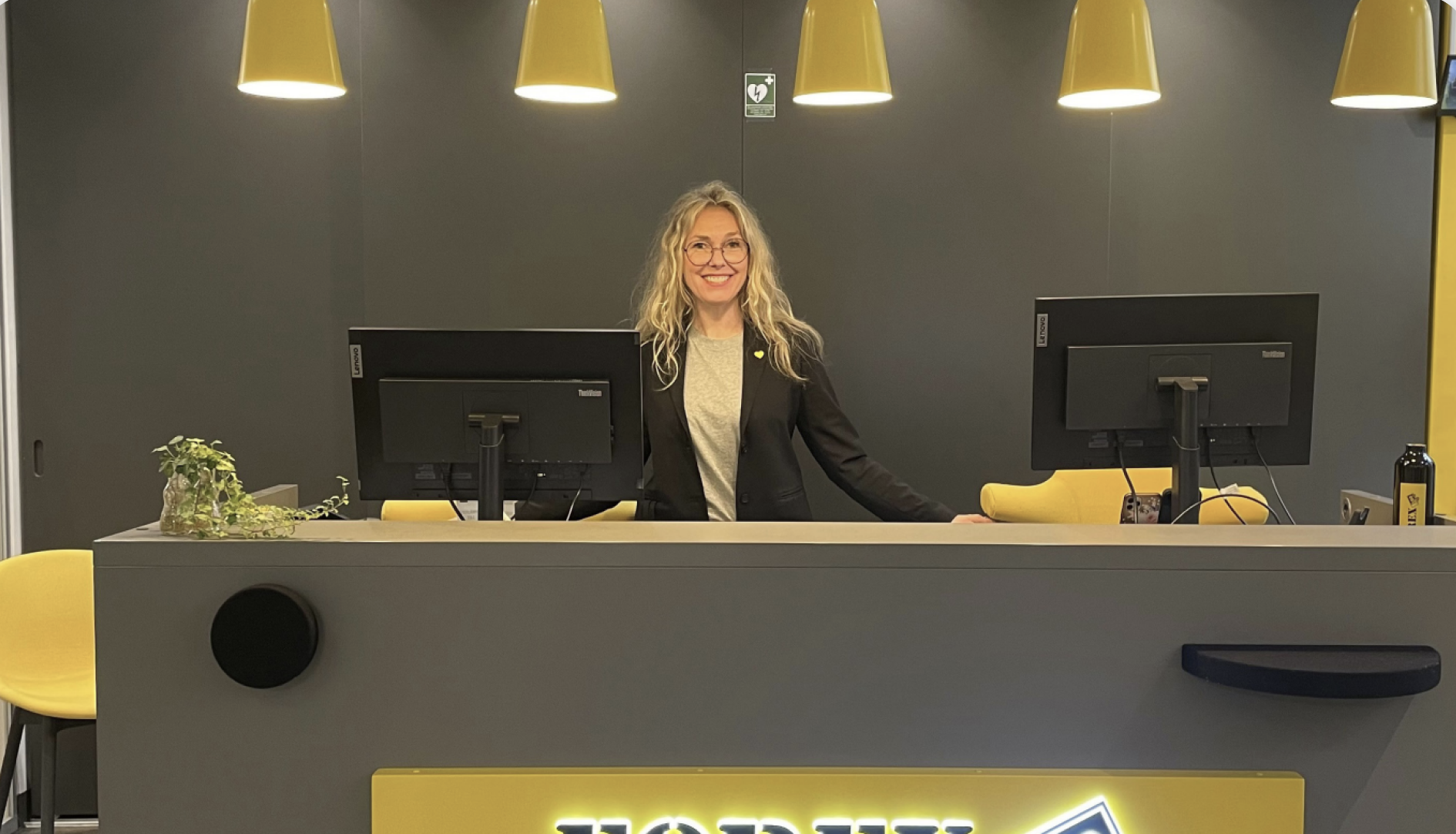 Receptionist standing at a modern office desk with dual monitors, surrounded by yellow decor and lighting.