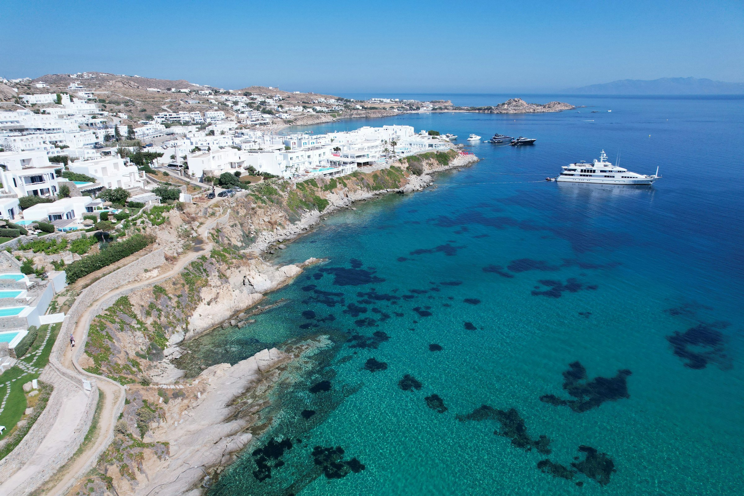Luftfoto av kusten på Mykonos, Grekland, med turkost hav, vita byggnader och båtar vid hamnen.