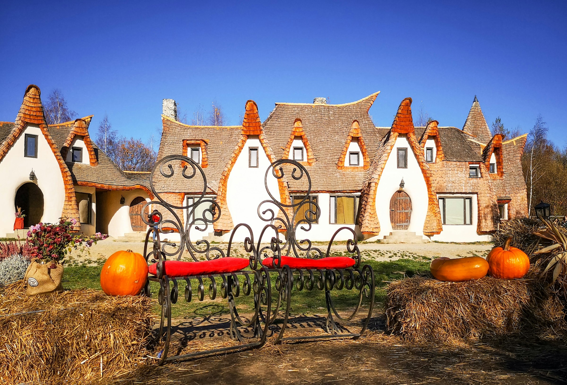 Sagolik arkitektur med spirade tak på ett ovanligt hus, smidesjärnsbänkar och höstliga pumpor framför byggnaden under en klarblå himmel.
