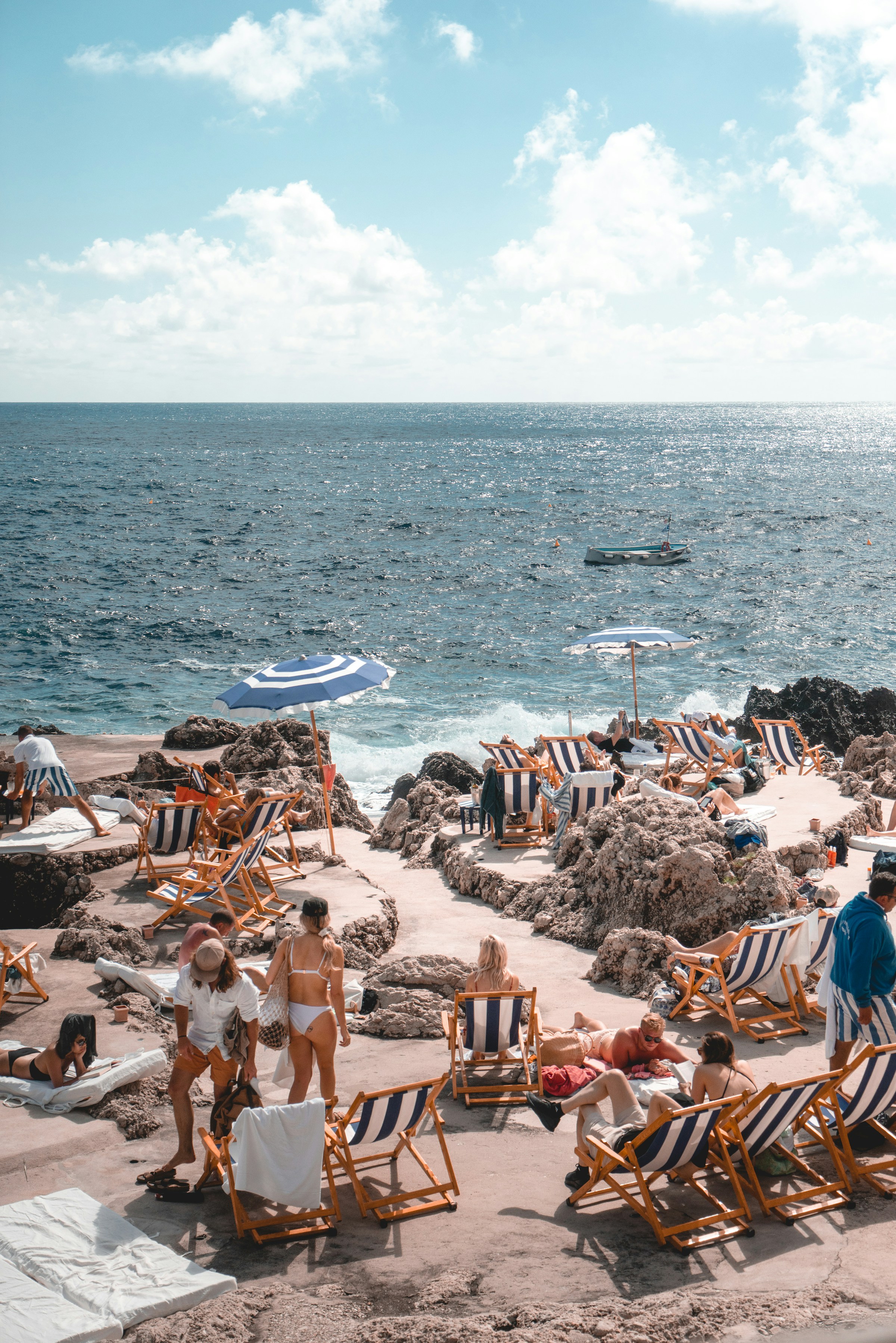Solstolar och parasoller vid klippig kust med turkost hav och badgäster som njuter av solsemester vid Medelhavet. Avkopplande strandmiljö i Sydeuropa.