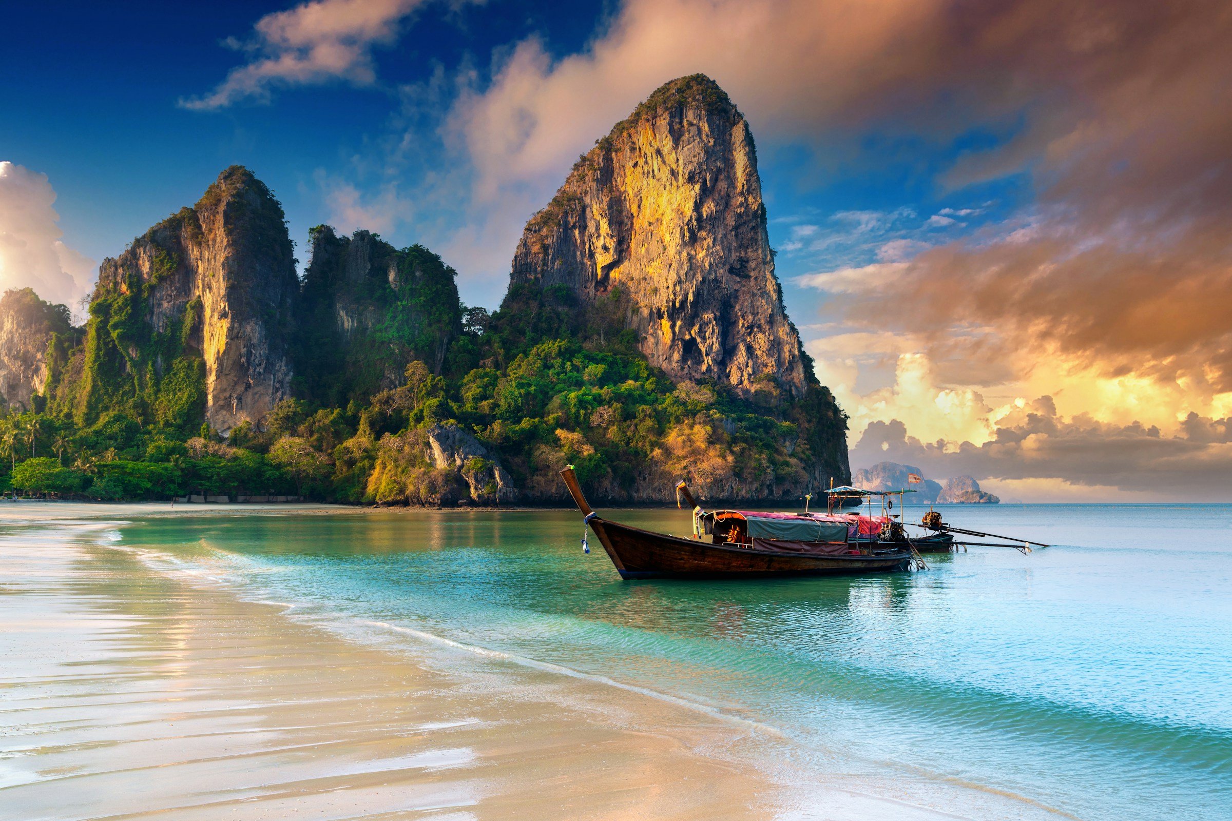 Best Trips Winter 2024 - Beach with traditional Thai longboat, crystal clear waters, and dramatic limestone cliffs at sunset in Krabi, Thailand.