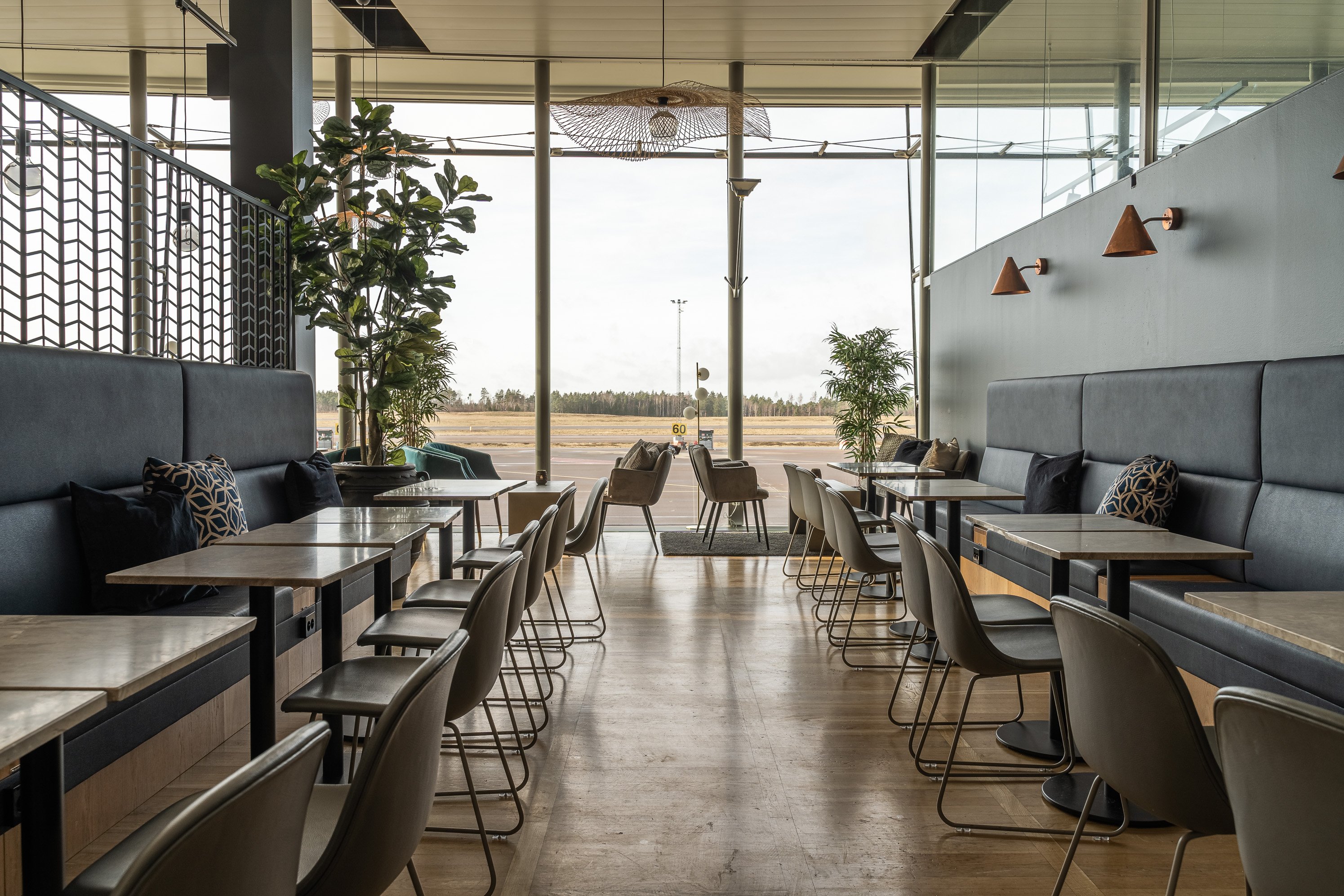Restaurangmiljö med rader av bord och stolar, stoppade bänkar längs väggarna och stora fönster med utsikt över ett öppet landskap.