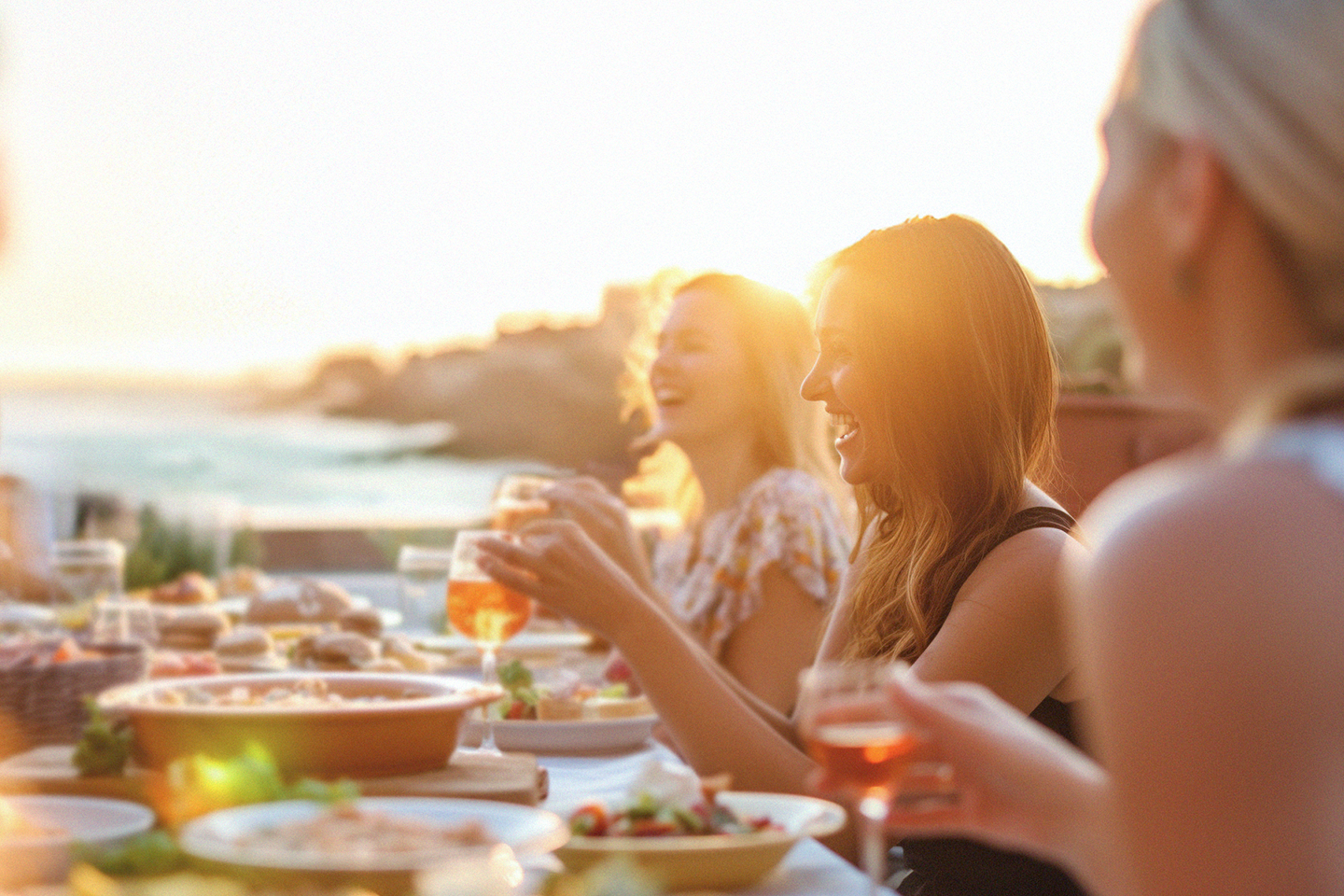 Människor njuter av en middag utomhus vid solnedgången, skrattar och håller glas med dryck.