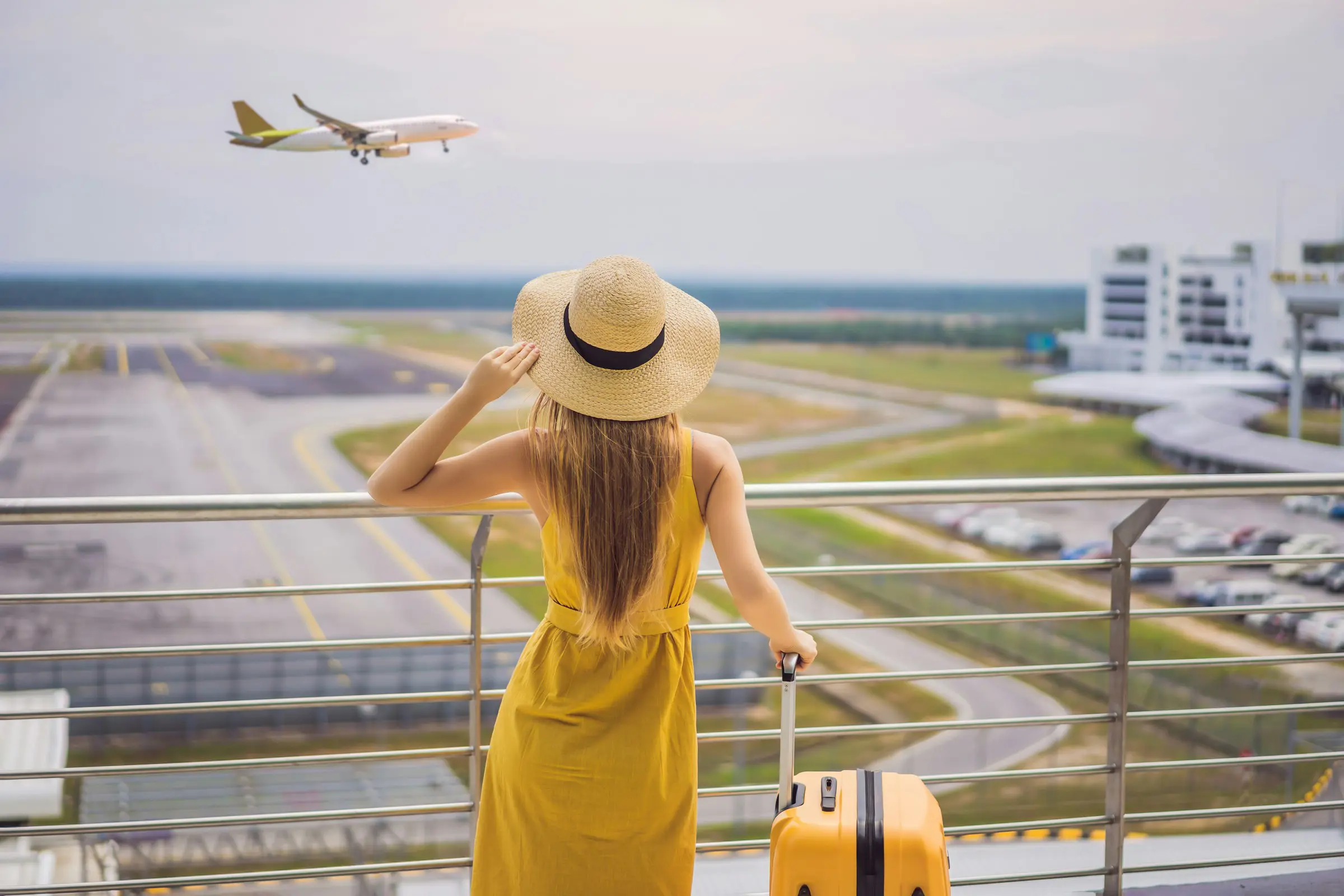 Flight search - Woman in yellow dress and straw hat watching airplane take off from airport observation deck, with yellow suitcase beside her.