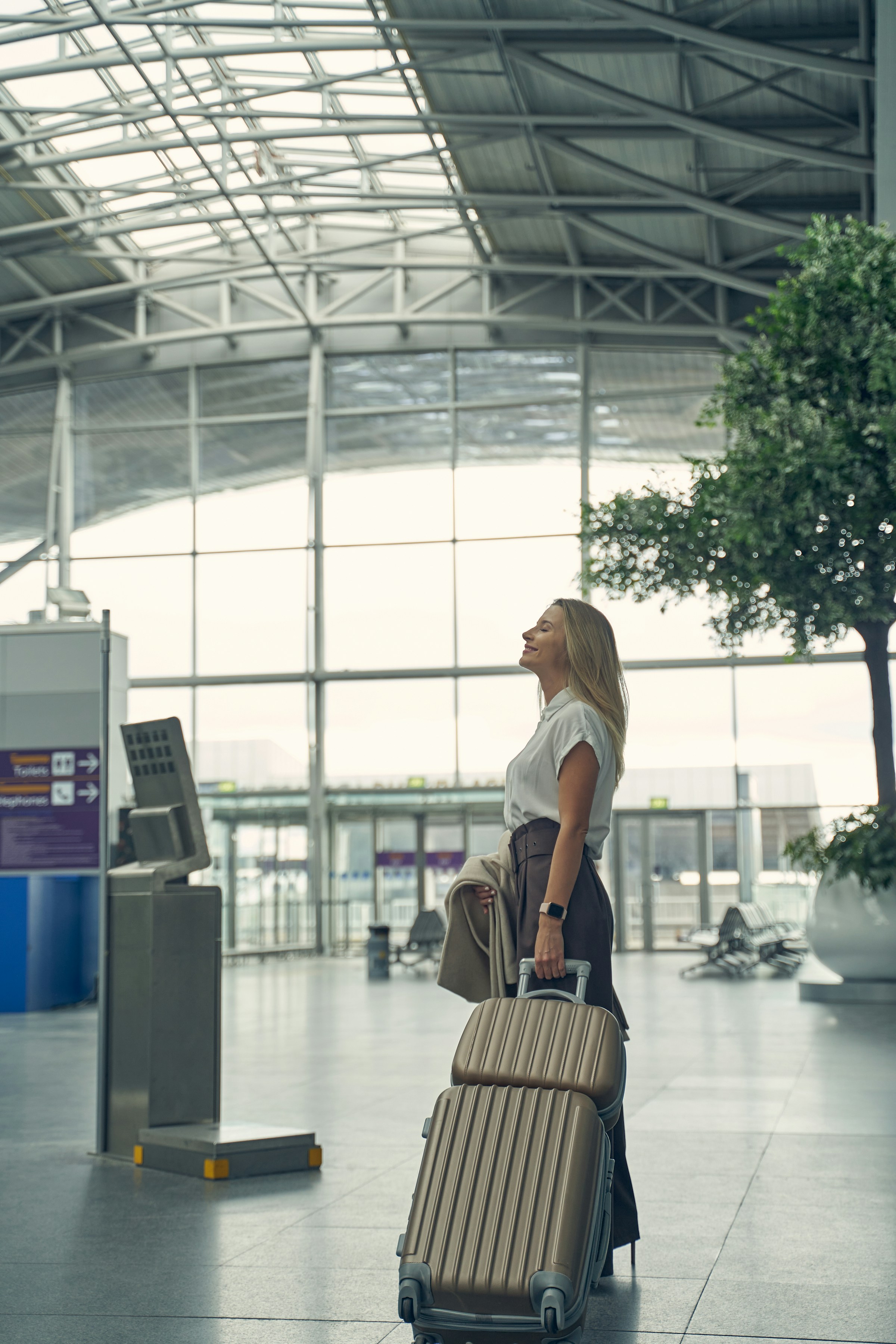 Woman standing in airport terminal with suitcase, looking upwards.