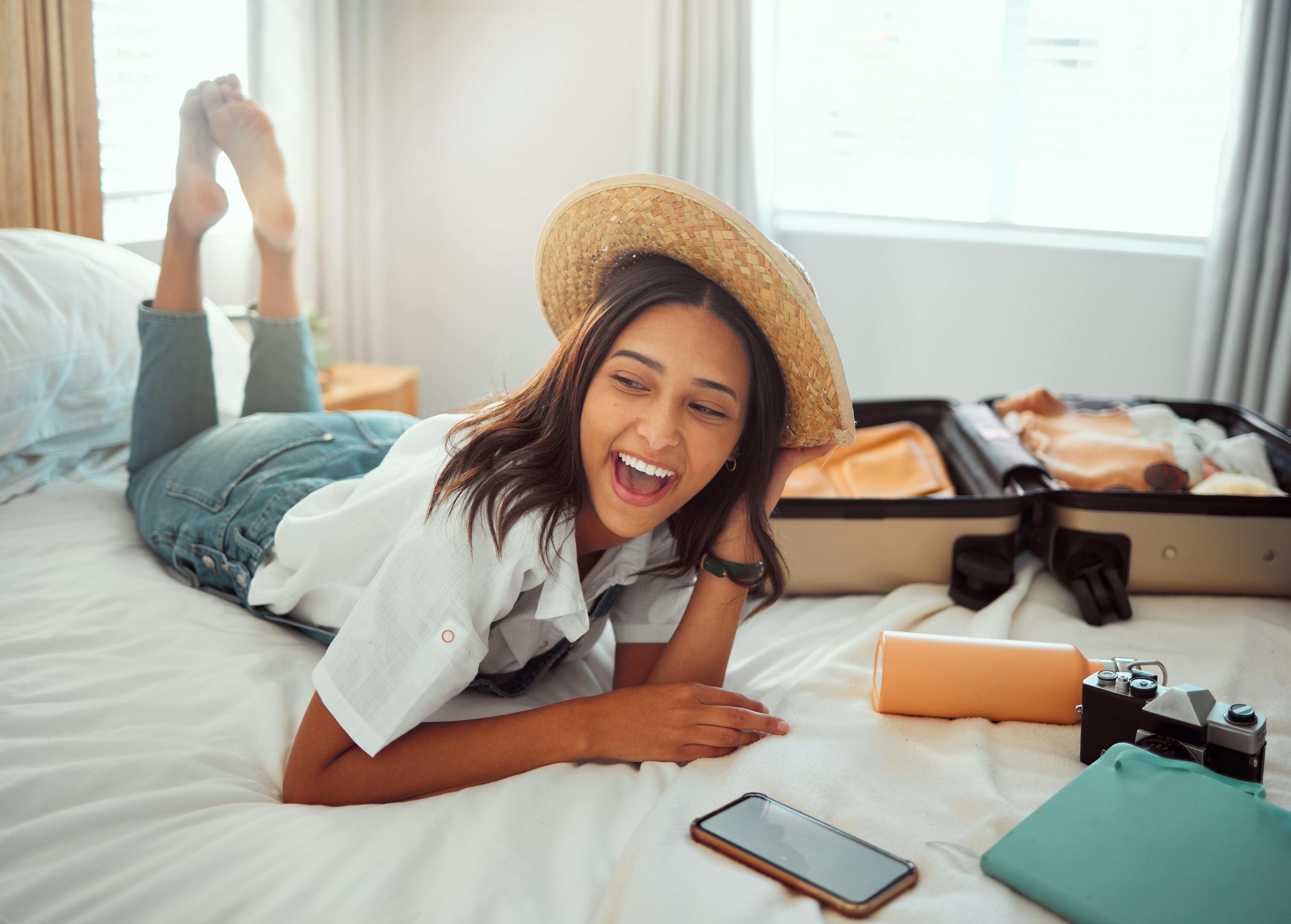 Woman in relaxed pose on bed with straw hat, smiling, next to open suitcase and smartphone, in a sunny room.