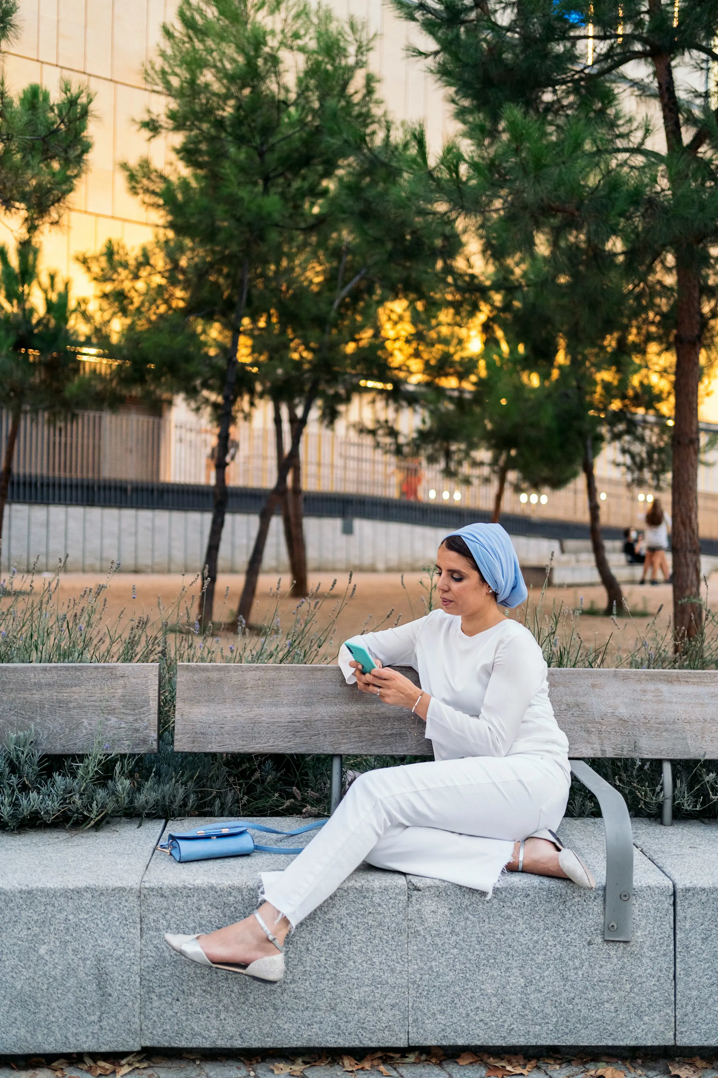 Woman in white outfit with a light blue headscarf sits on a bench using her phone, surrounded by trees and an urban background.