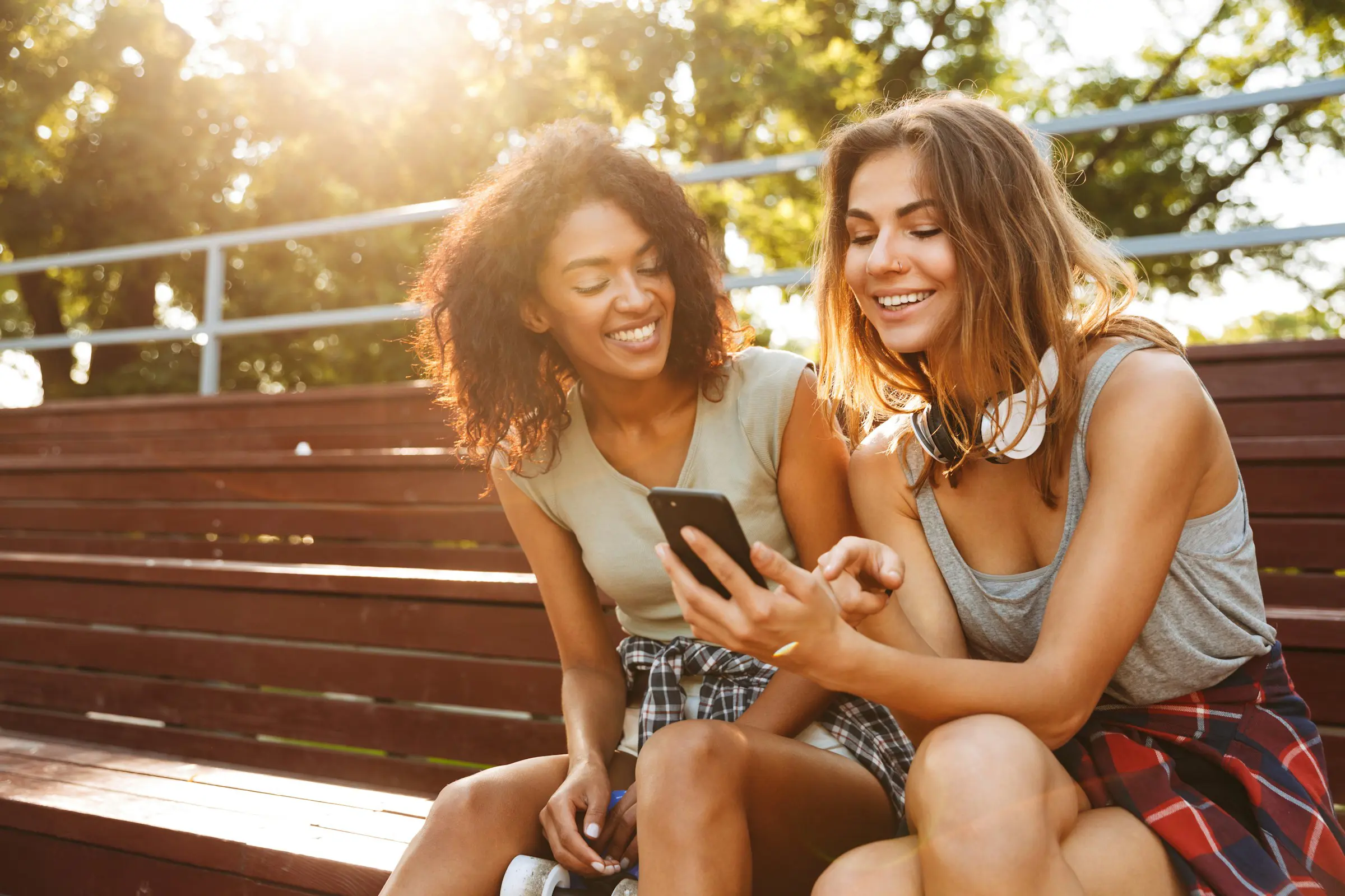 Två glada unga kvinnor som tittar på smartphone och ler utomhus i parken.