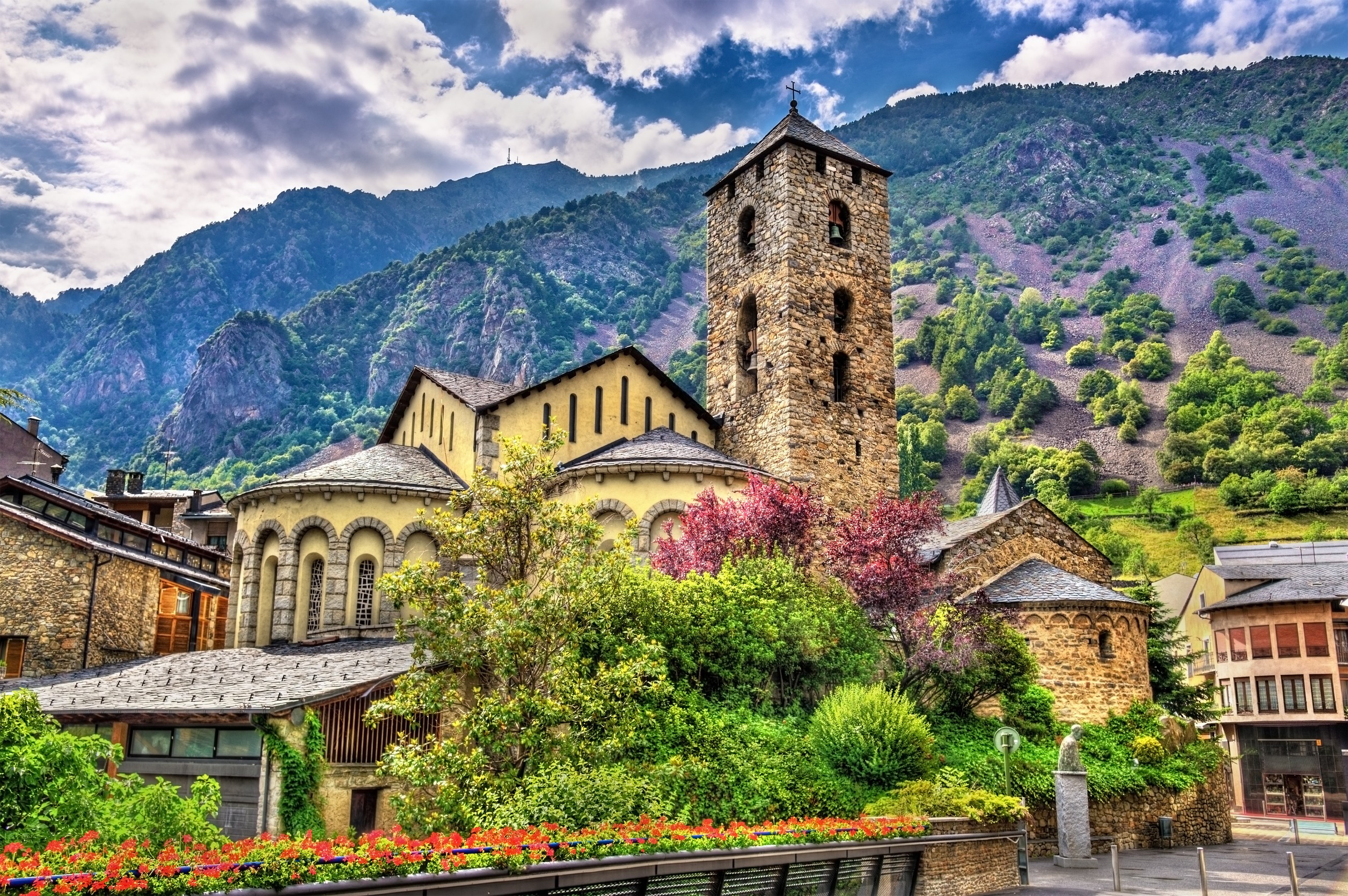 Romanesque church in Andorra la Vella surrounded by vibrant greenery and colorful flowers, set against a backdrop of majestic mountains under a partly cloudy sky.