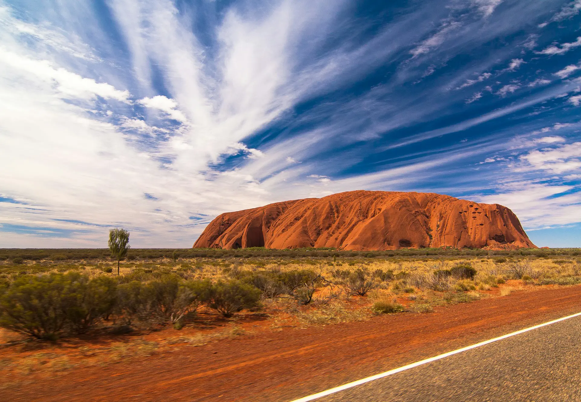 Uluru, a large sandstone monolith in the Australian Outback, under a vibrant blue sky with streaked clouds and red desert landscape in the foreground.