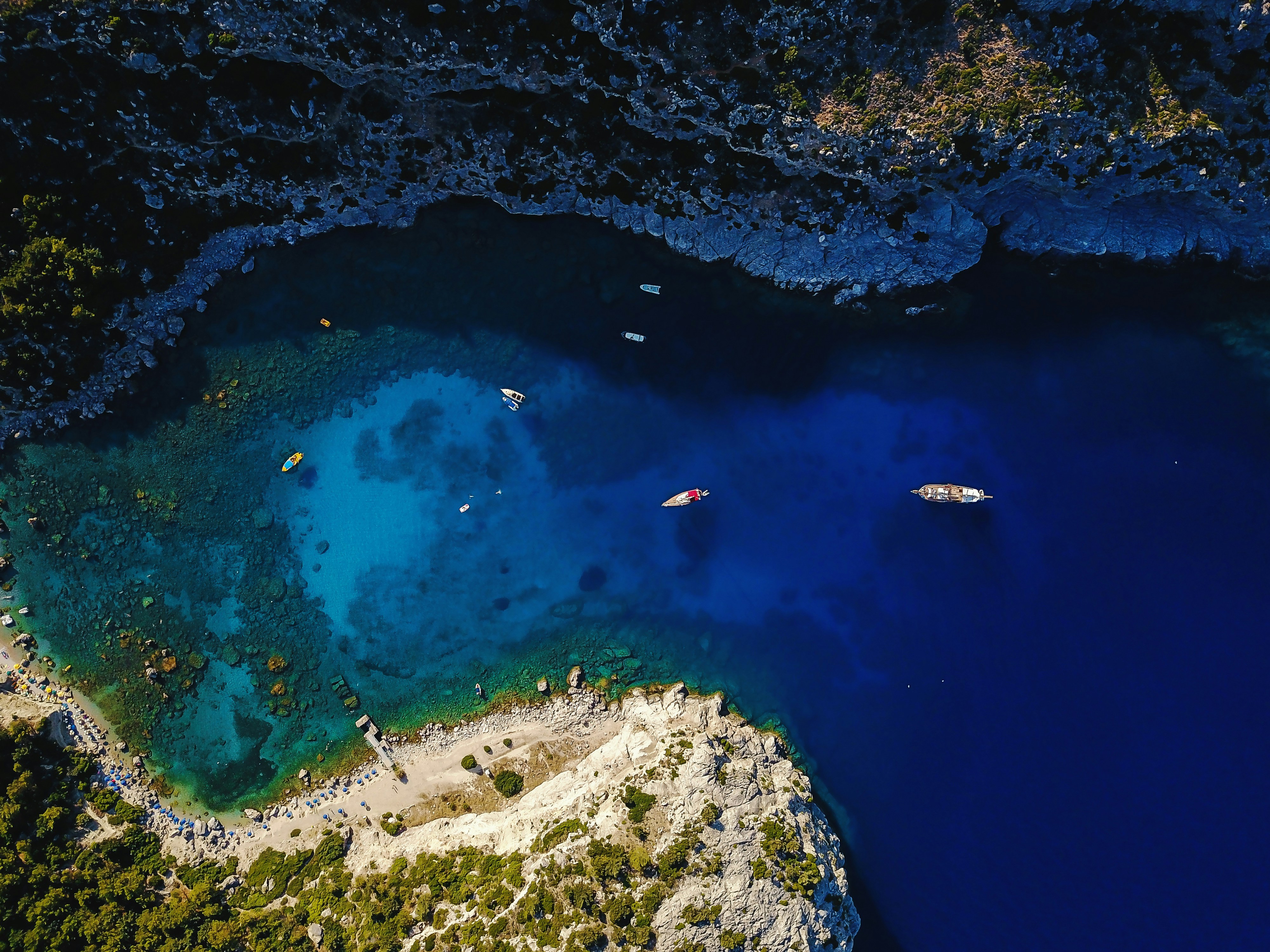 Aerial view of a serene bay with vibrant blue waters, surrounded by rocky cliffs and lush greenery. Small boats are scattered across the water.