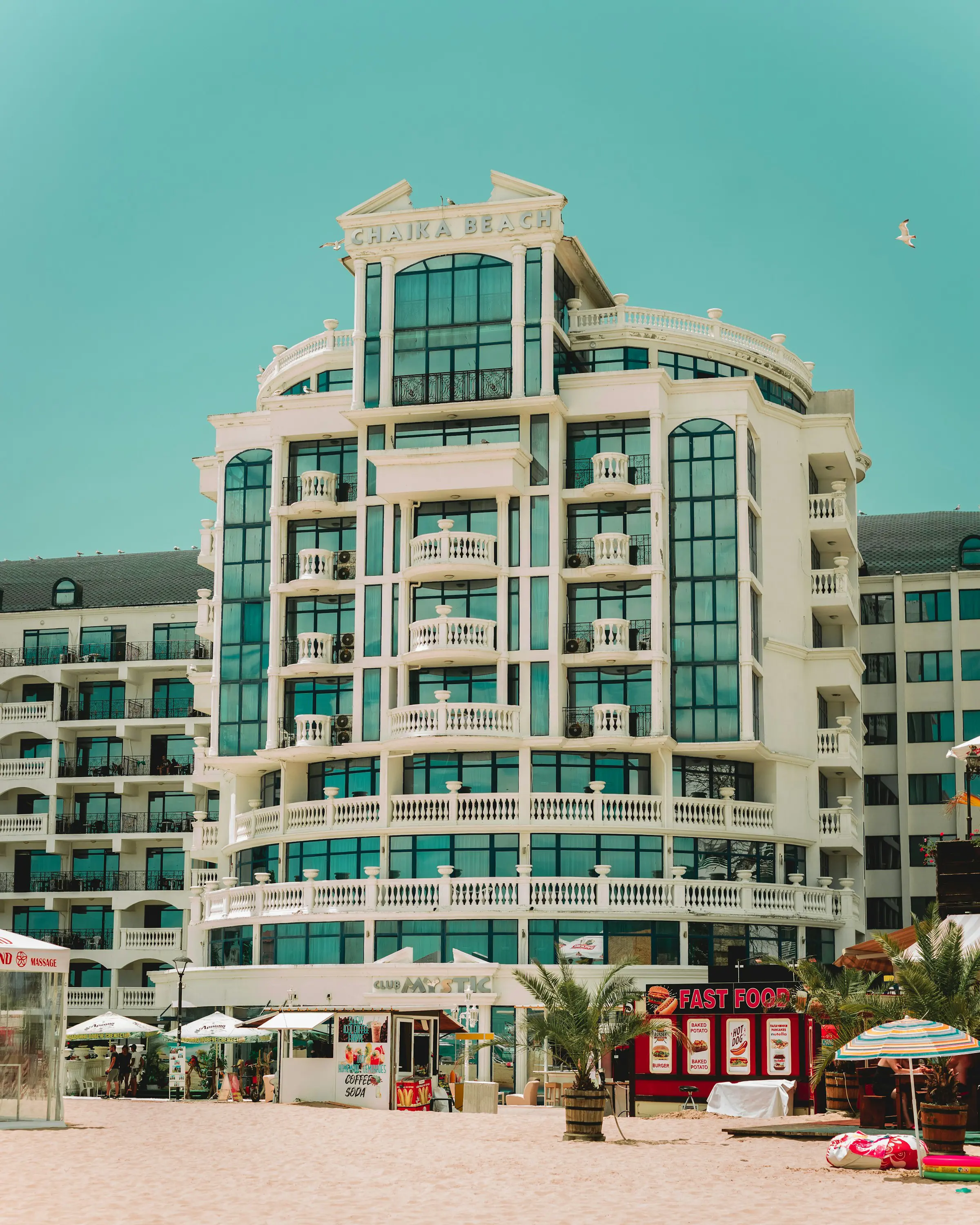 Modern beachfront hotel with glass balconies and resort amenities, set against a clear blue sky and sandy beach, featuring nearby food stalls and palm trees.