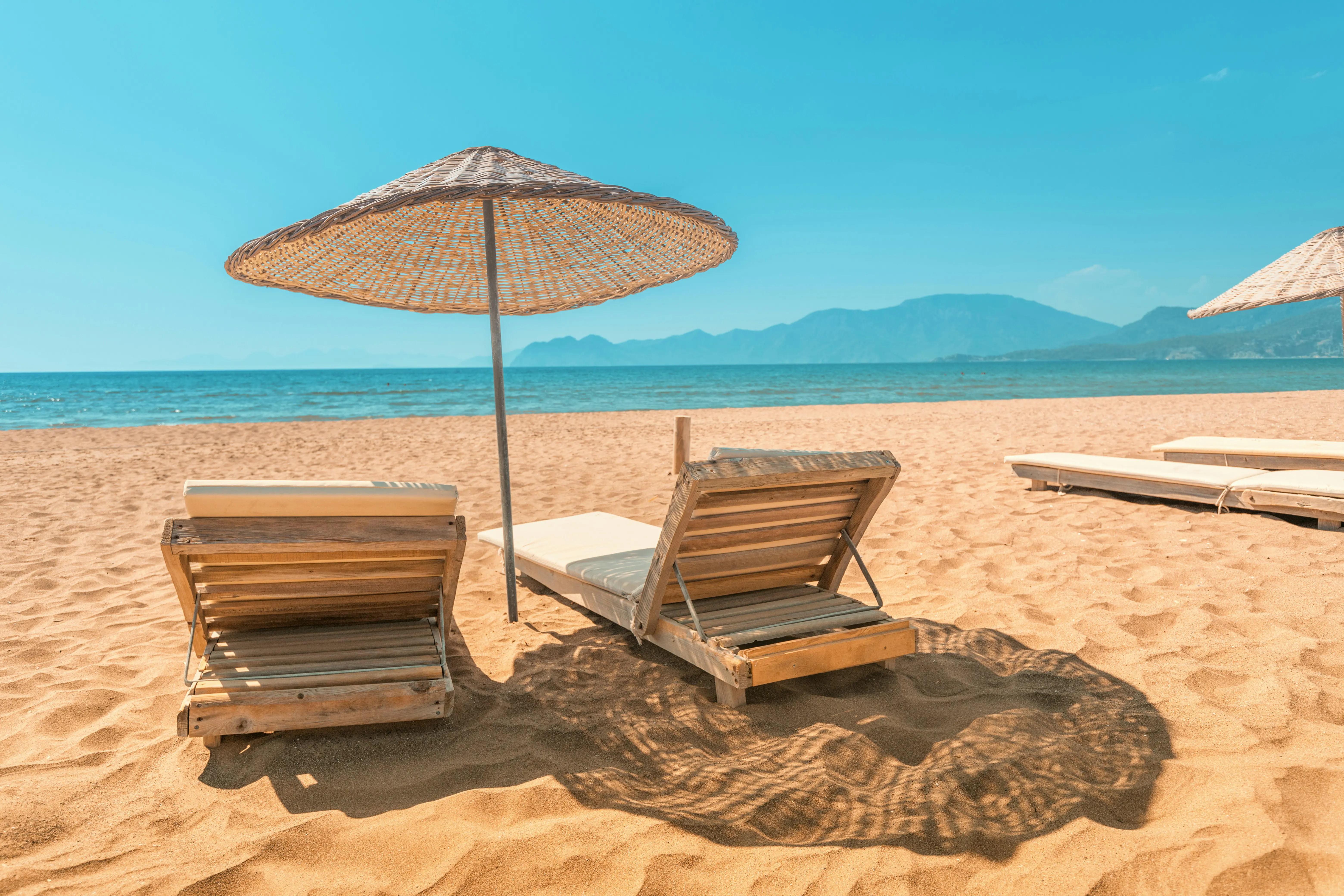 Sandy beach with two wooden lounge chairs and a straw umbrella, overlooking a serene ocean and distant mountains under a clear blue sky.