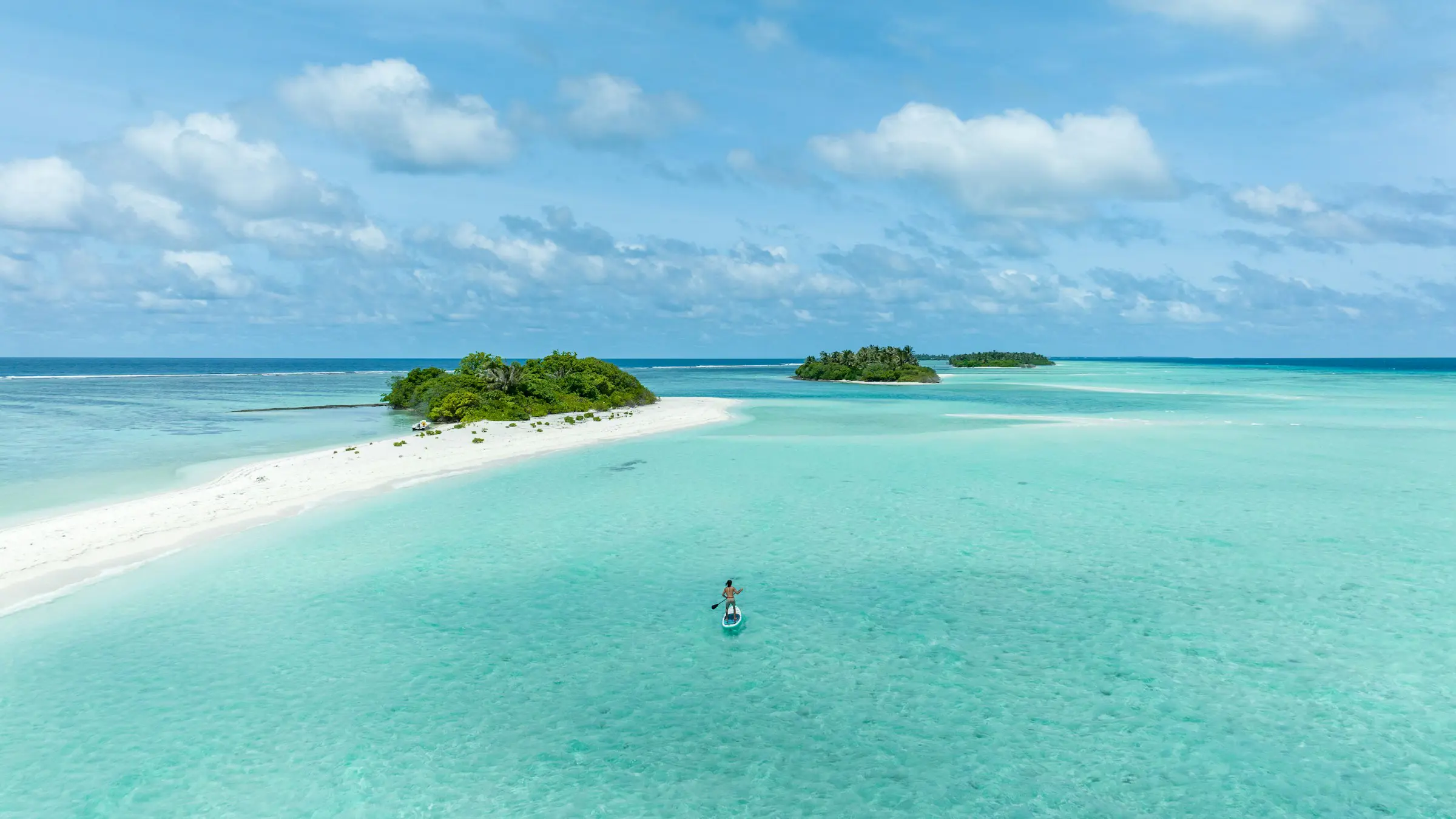 A person paddleboarding in clear turquoise water near small tropical islands with lush greenery under a blue sky.