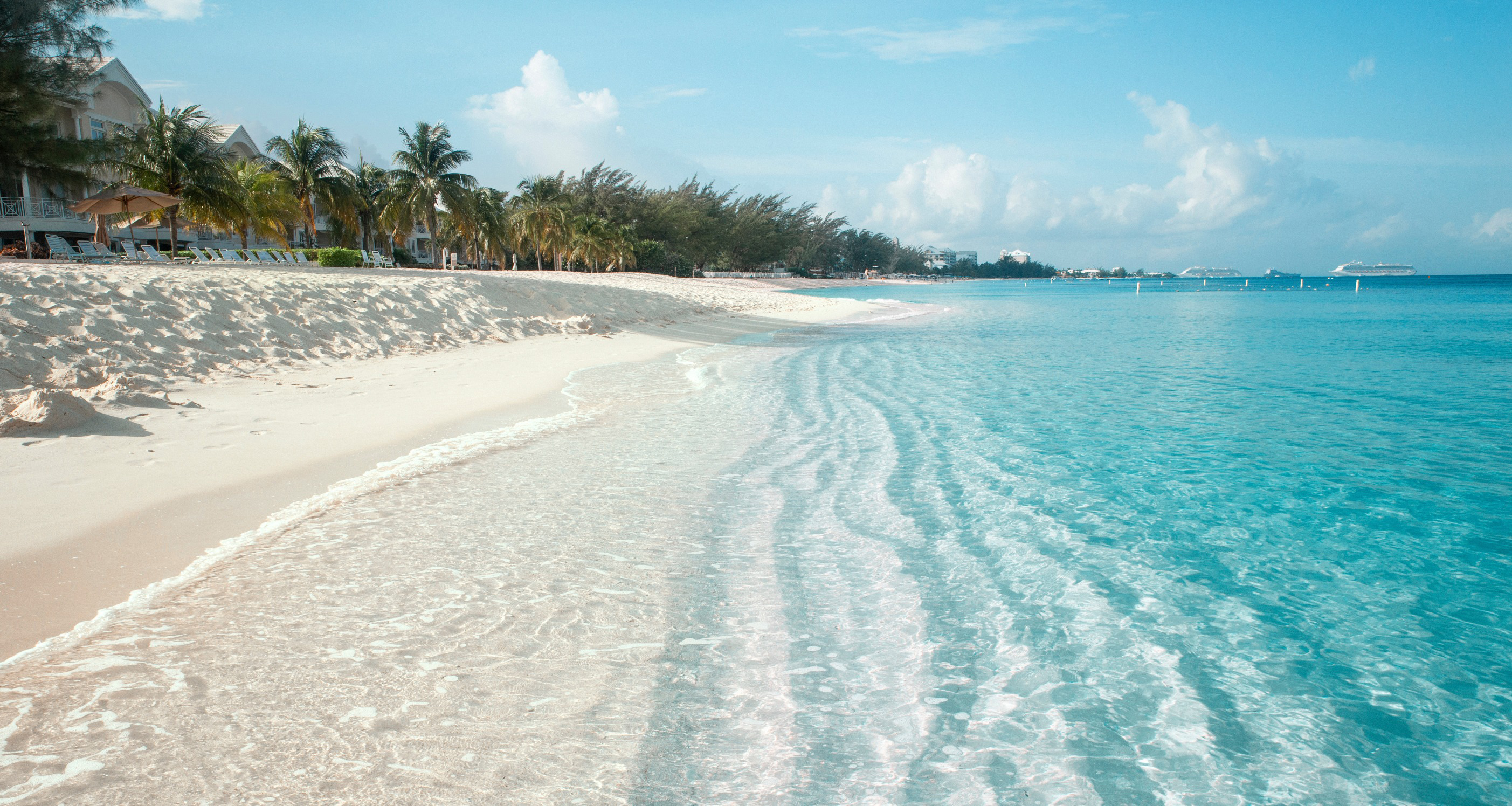 Travel to Cayman Islands - Pristine white sandy beach with clear turquoise water under a blue sky, lined with palm trees and distant buildings.