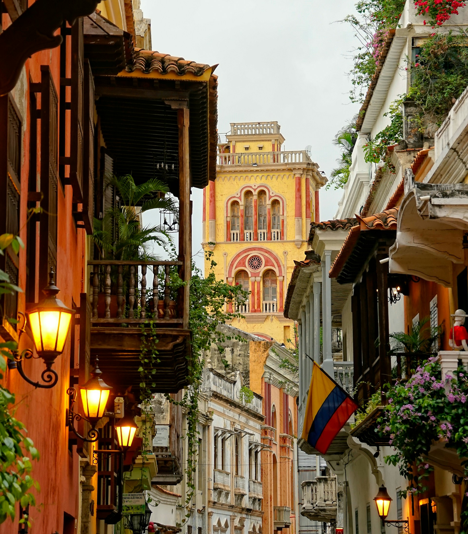 Charming street view in Cartagena, Colombia, featuring colonial architecture, vibrant building facades, hanging plants, lanterns, and the Colombian flag.