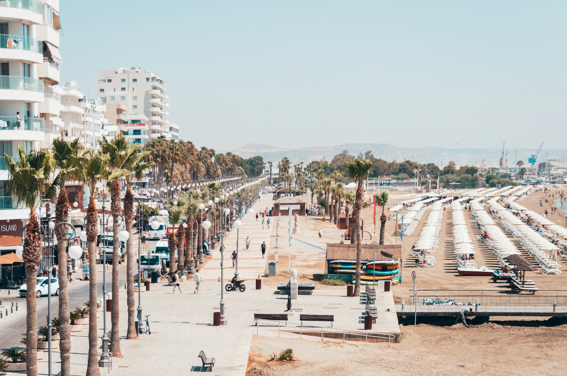 Beach promenade with palm trees and high-rise buildings in Larnaca, Cyprus, featuring sunbathers under umbrellas along a sandy beach.
