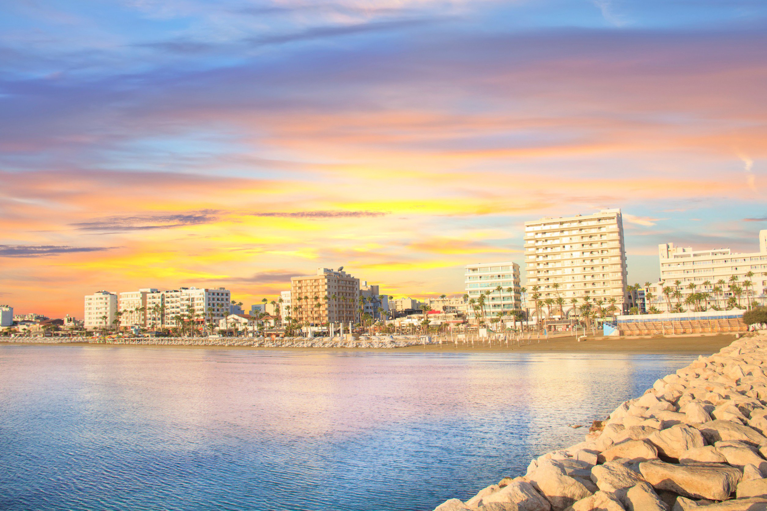 Travel to Larnaca - Sunset over the coastline with high-rise buildings, viewed from a rocky shoreline, enhancing the urban waterfront and vibrant sky.