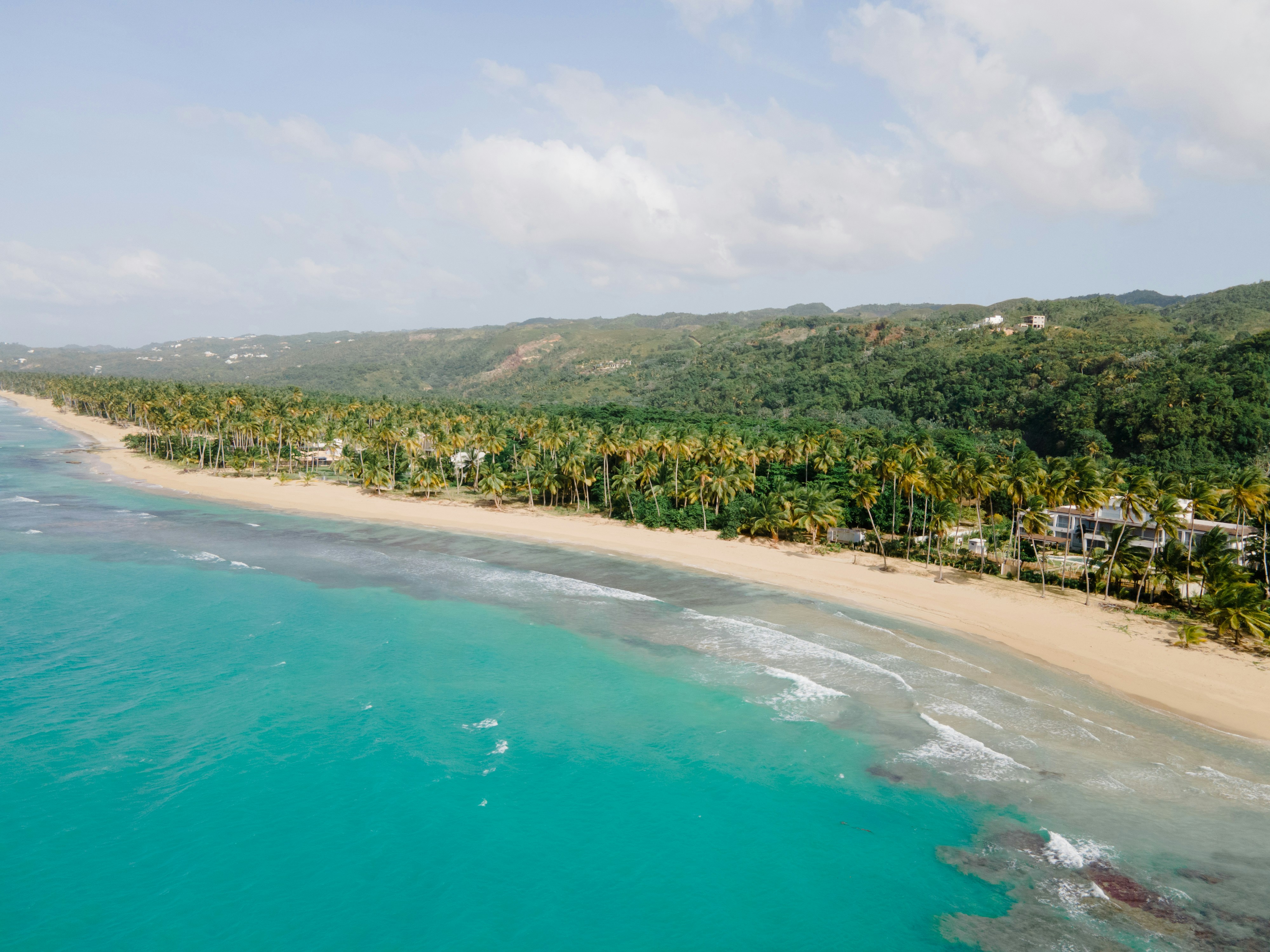 Aerial view of a tropical beach with turquoise waters, sandy shoreline, and lush green palm trees, set against a backdrop of green hills under a partly cloudy sky.