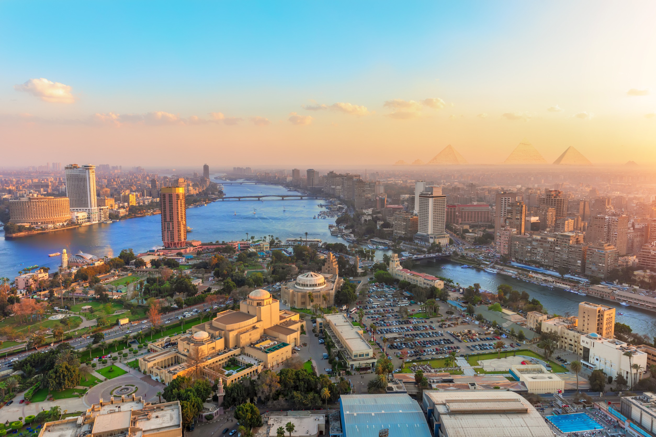 Travel to Cairo - view of Cairo cityscape with the Nile River flowing through, surrounded by urban buildings and greenery at sunset, with the Giza Pyramids visible.