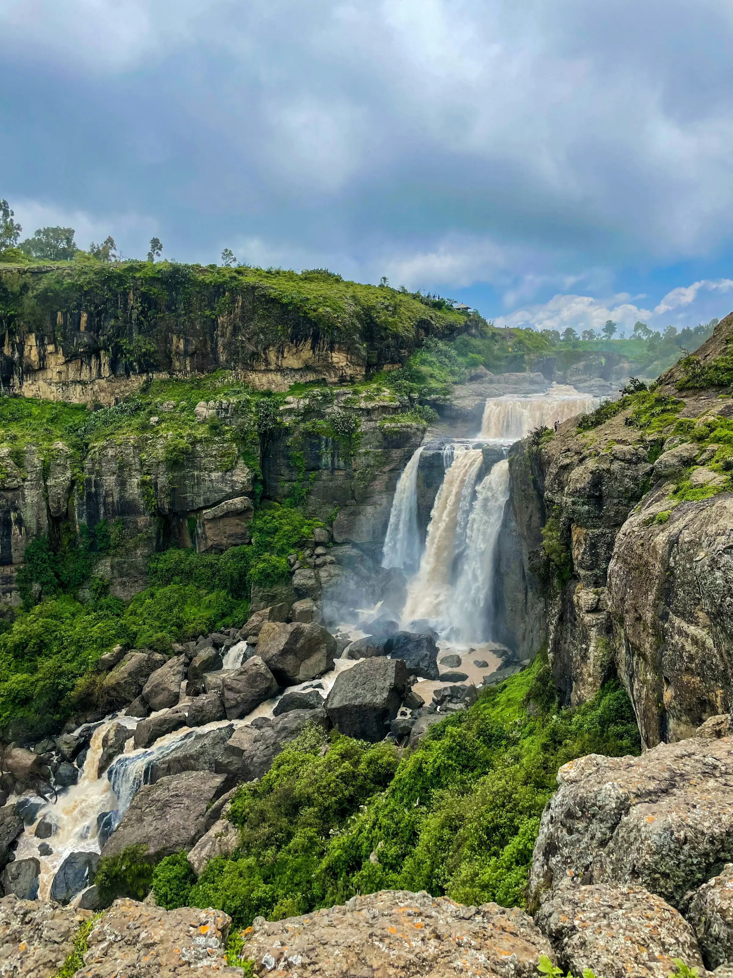 A scenic waterfall cascading over a cliff surrounded by lush greenery and rocky terrain under a cloudy sky.