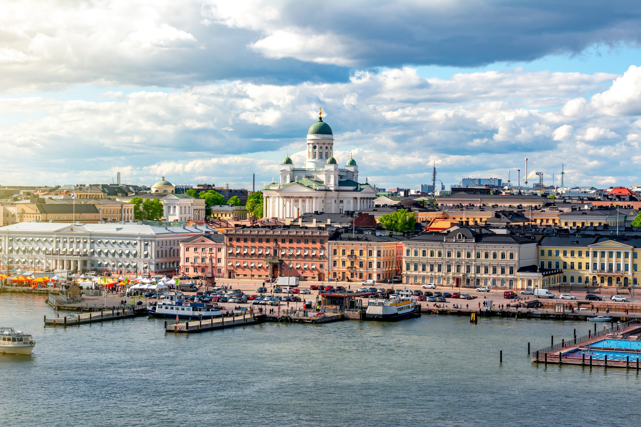 Travel to Helsinki - Scenic view of waterfront with colorful buildings and Helsinki Cathedral in the background under a cloudy sky.