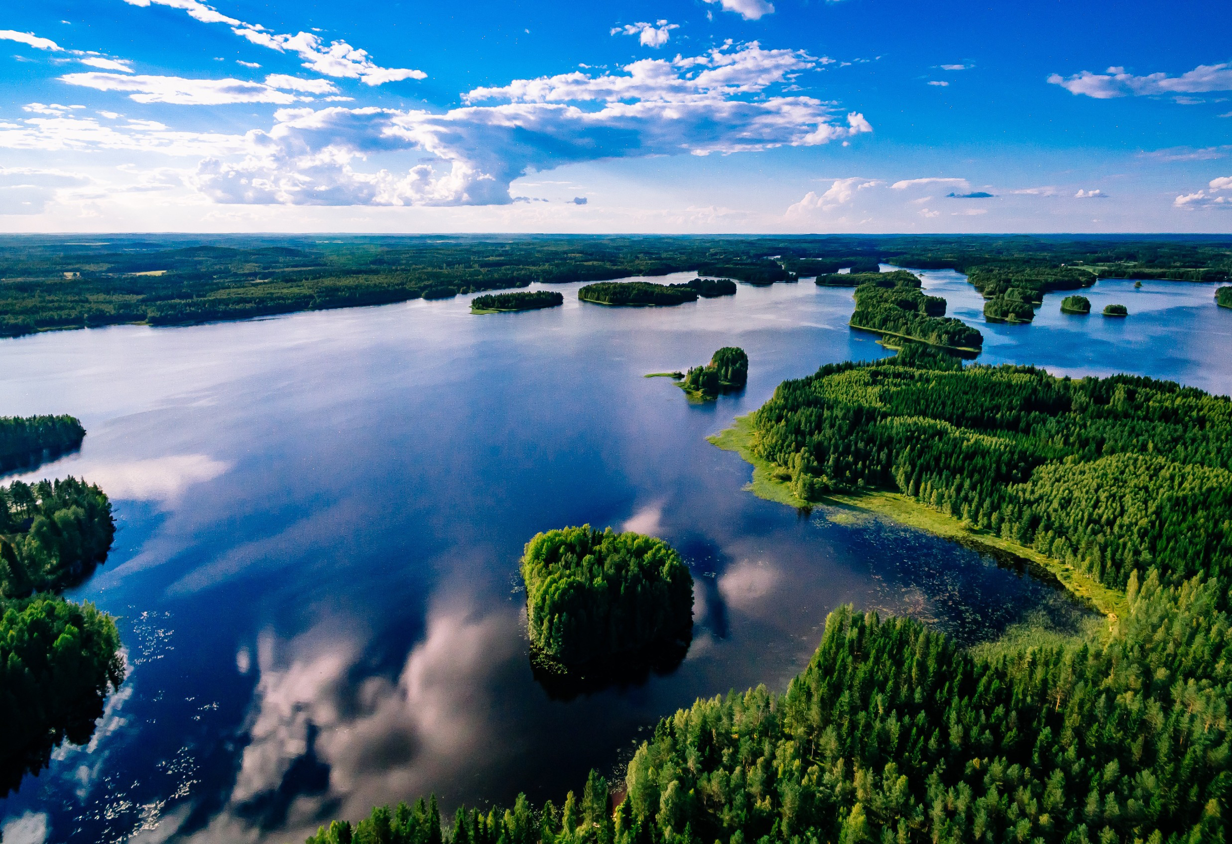 Travel to Finland - Aerial view of a scenic Finnish lake landscape with lush green islands, reflecting a partly cloudy sky over calm blue waters.