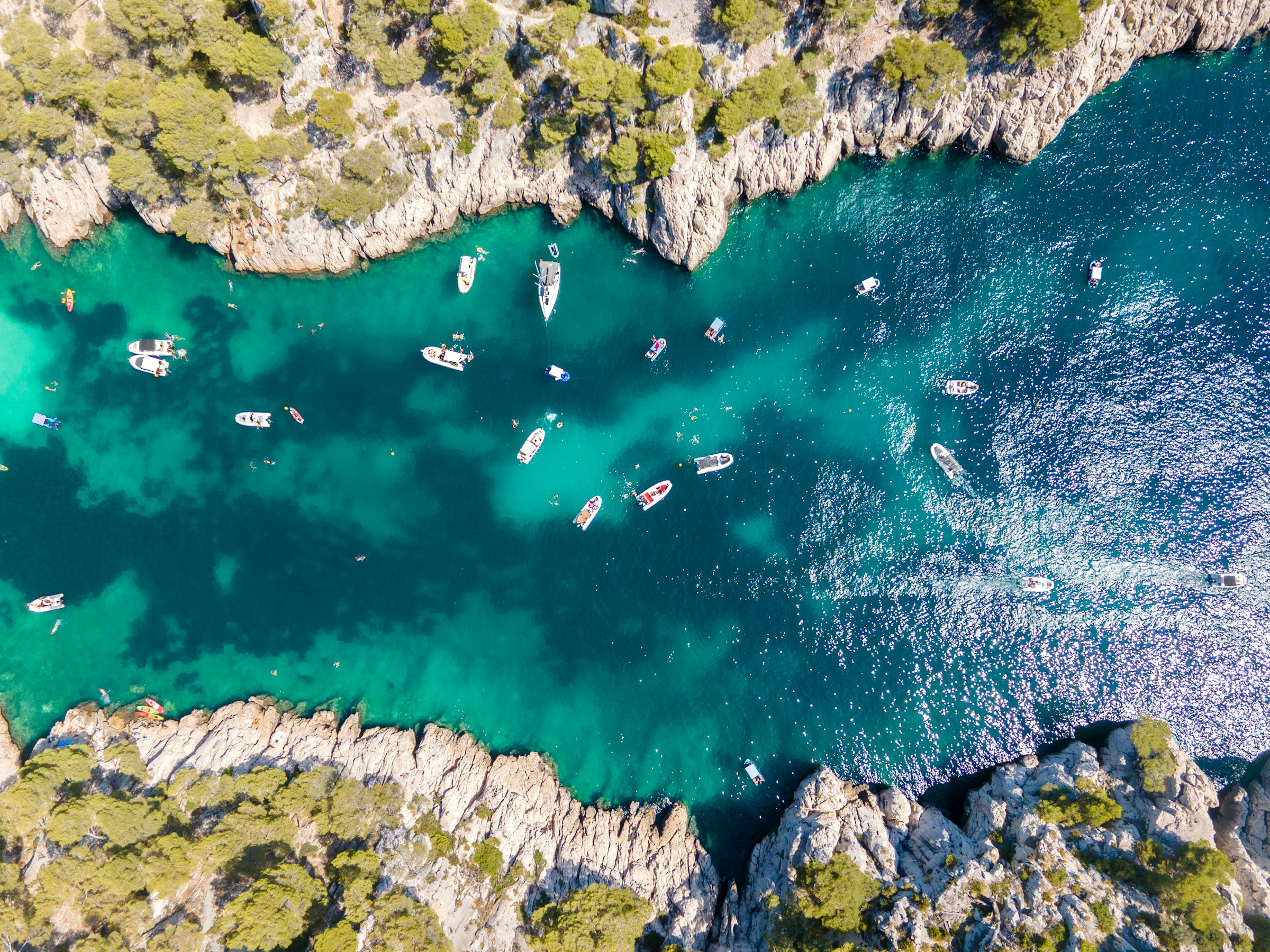 Aerial view of turquoise water surrounded by rocky cliffs with scattered boats and kayaks. Lush green trees line the coastline, creating a stunning natural landscape.