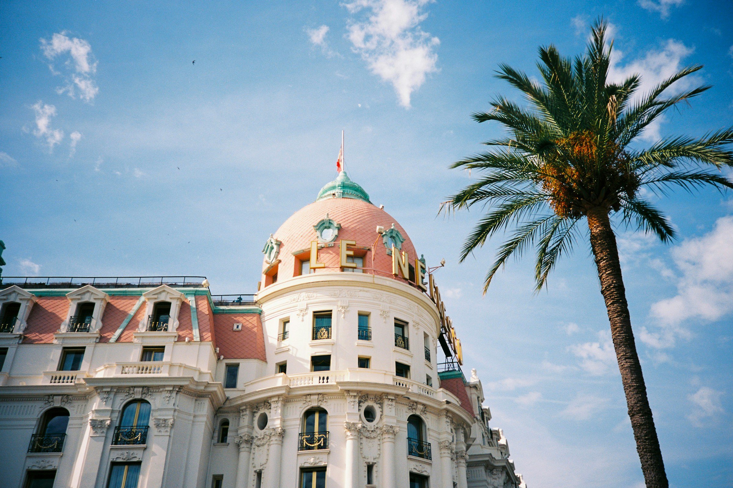 Historic hotel with ornate architecture and pink dome under a bright blue sky, featuring a tall palm tree beside it.