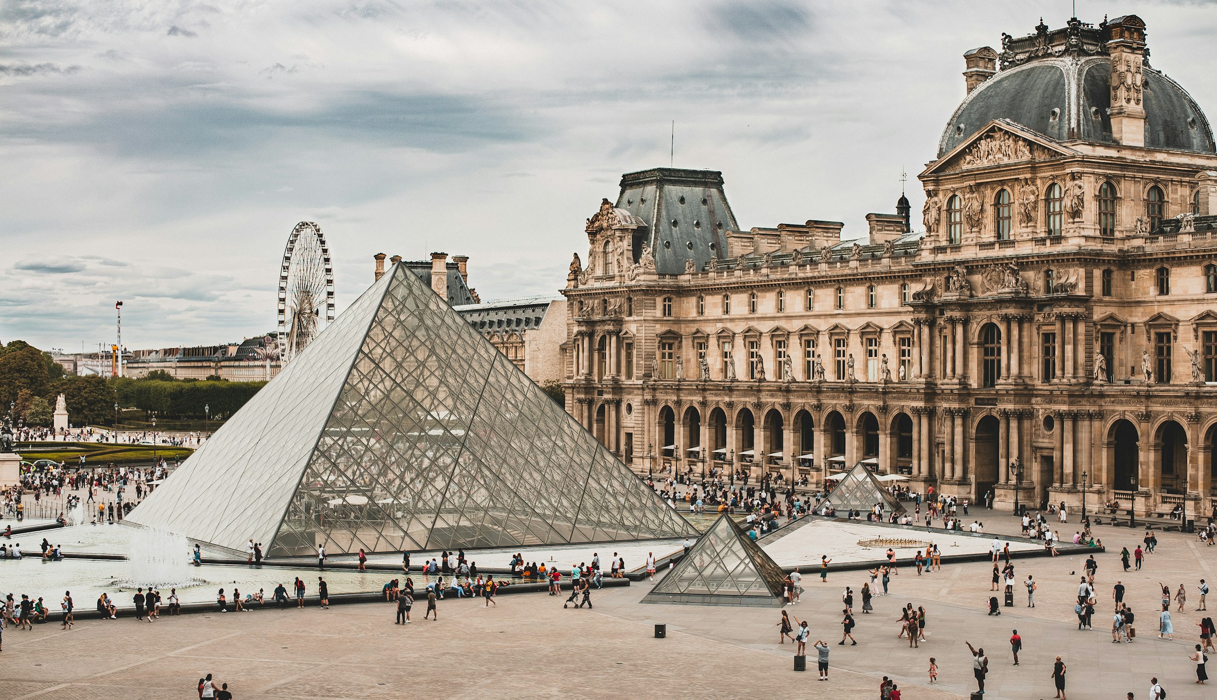 Wide view of the Louvre Museum with the iconic glass pyramid entrance in Paris, bustling with tourists.