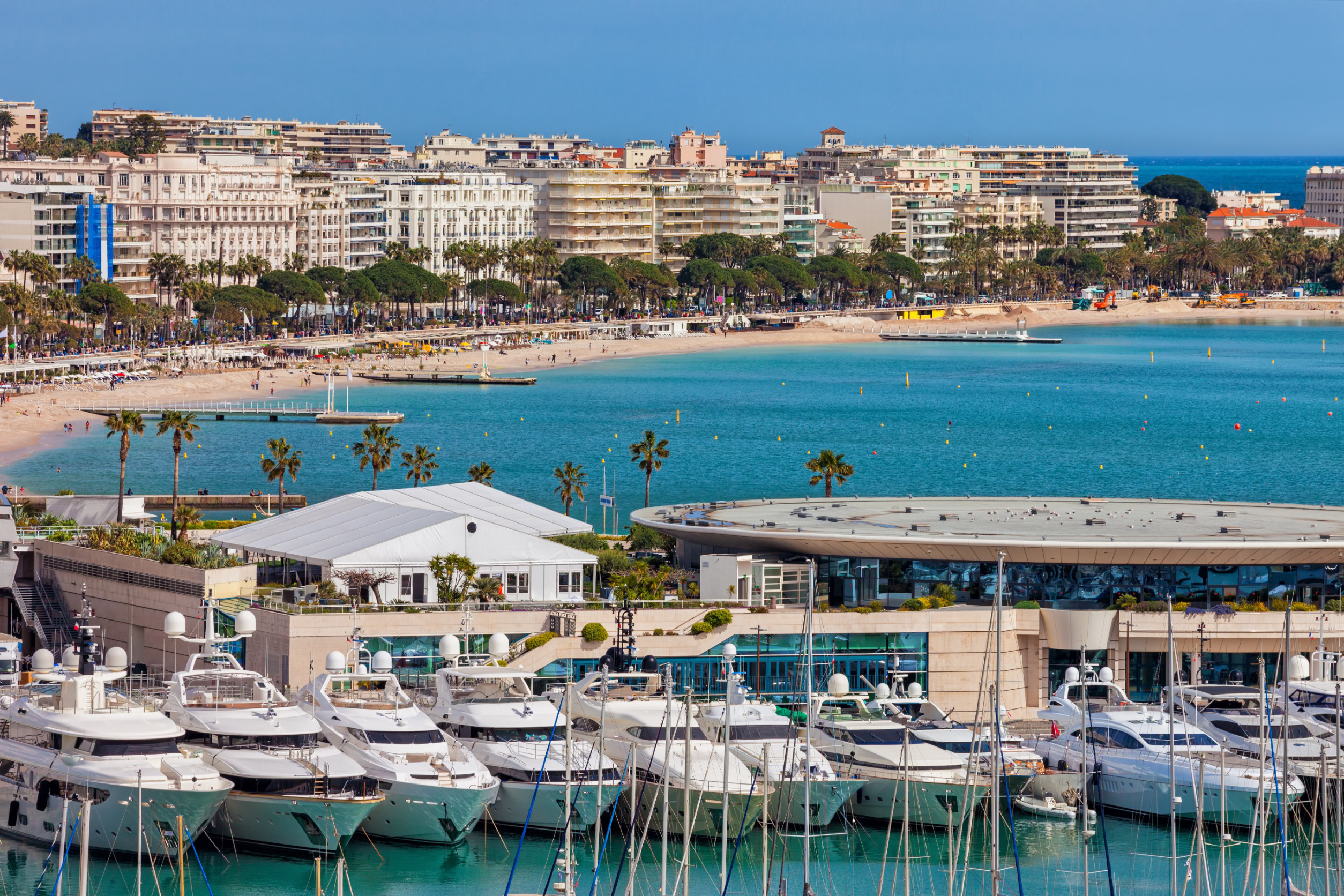 Travel to Cannes - Cannes Marina with luxury yachts, turquoise water, sandy beach, and urban buildings in the background.