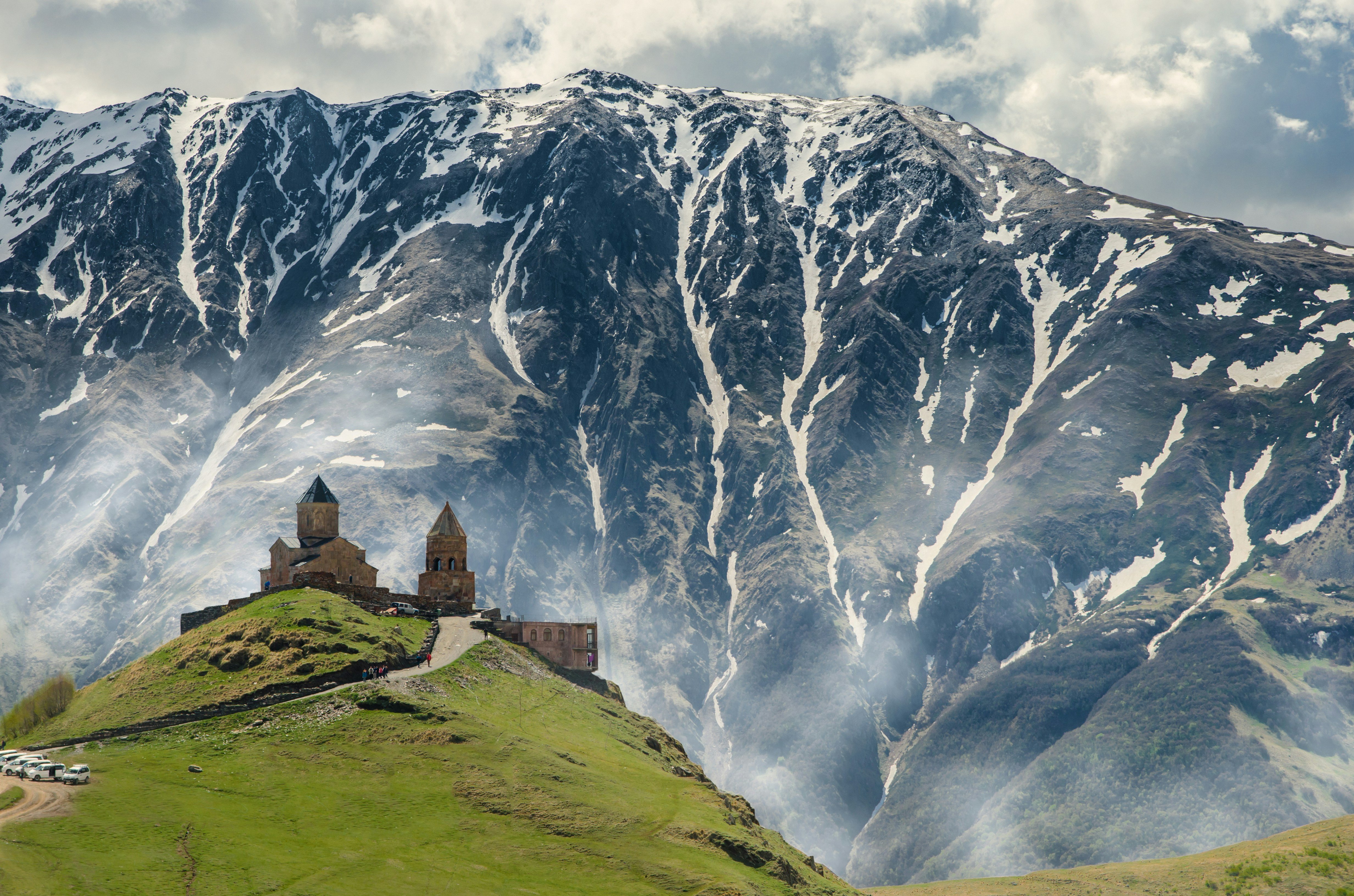 Travel to Georgia - Church on a green hill in front of dramatic snow-capped mountains.