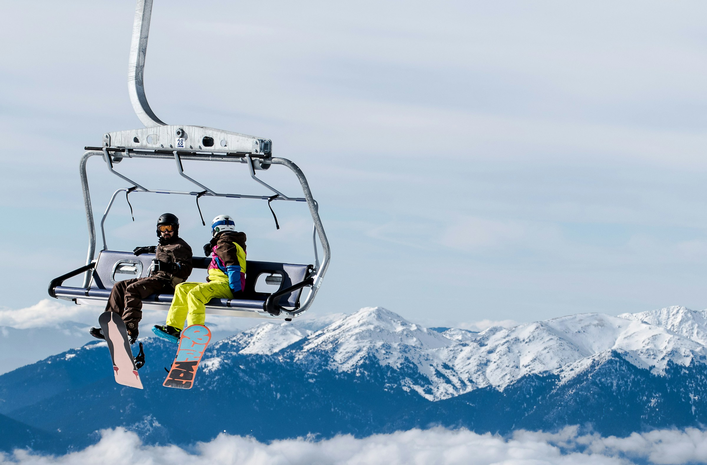Two snowboarders on a chairlift with snow-capped mountains in the background, clear sky.