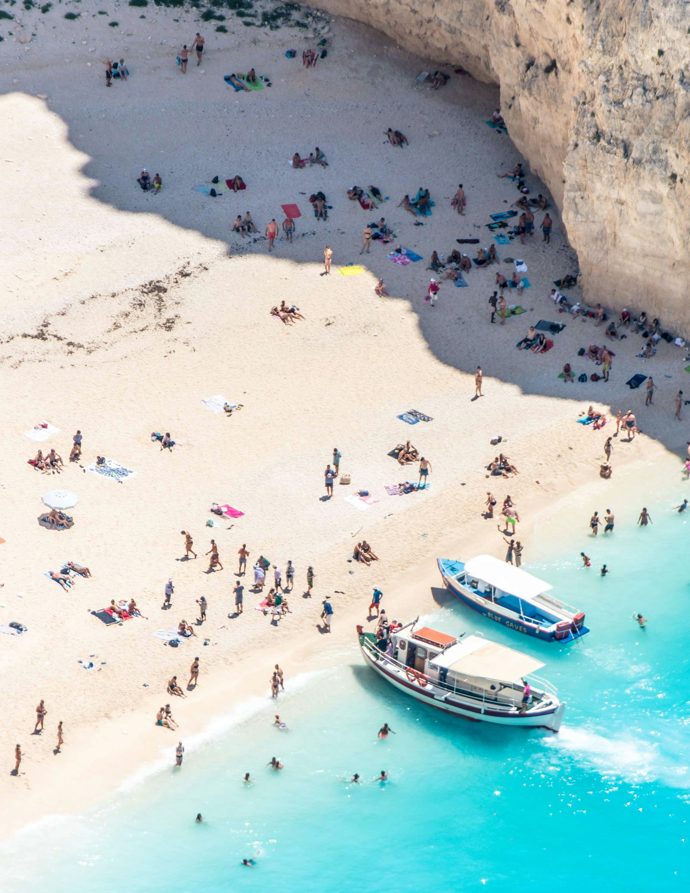 View of a crowded beach with people relaxing and swimming, near turquoise waters and rocky cliffs. Two boats are docked along the shore.