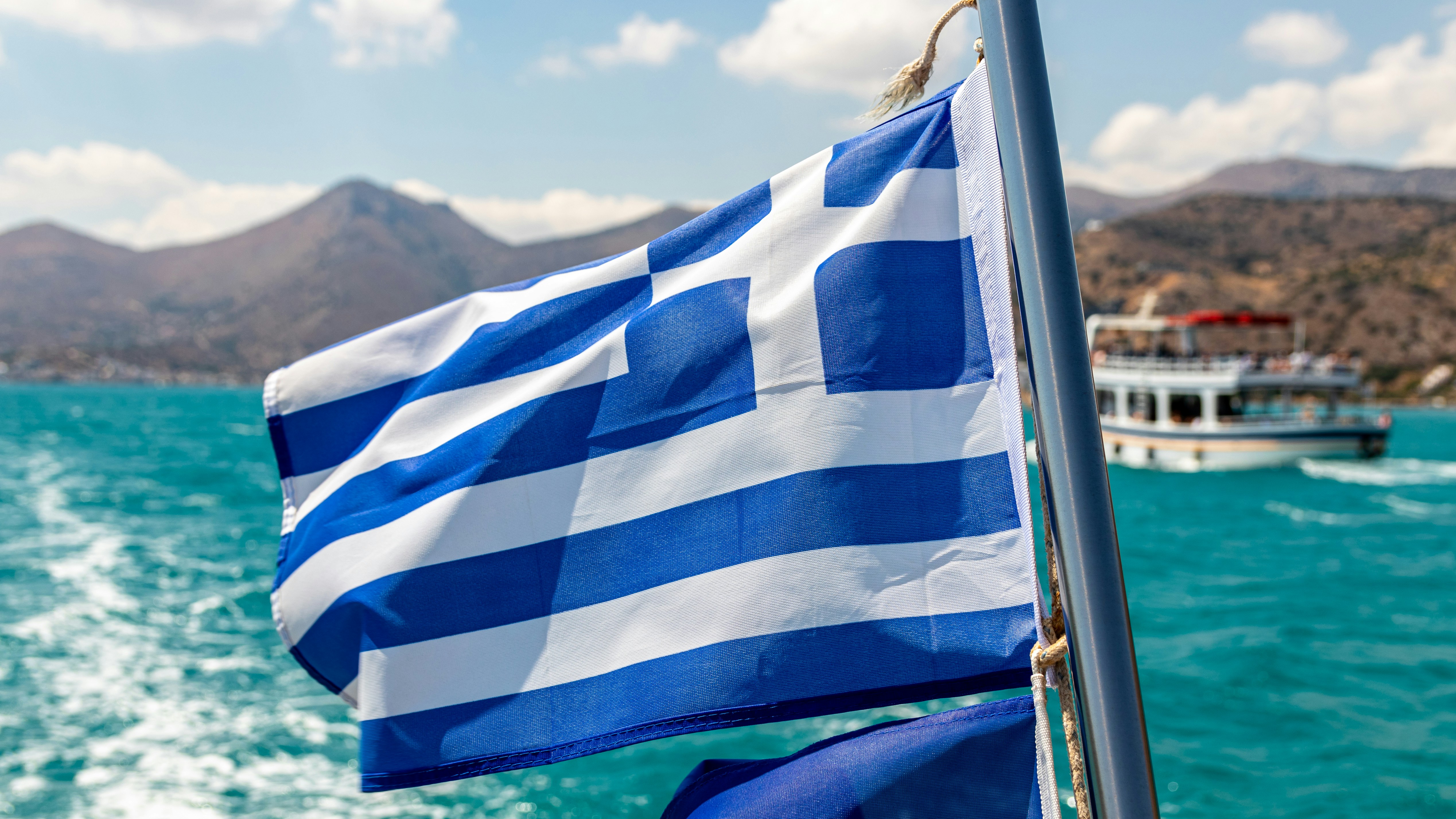 Greek flag waving on a boat with turquoise sea and distant mountains in Samos.