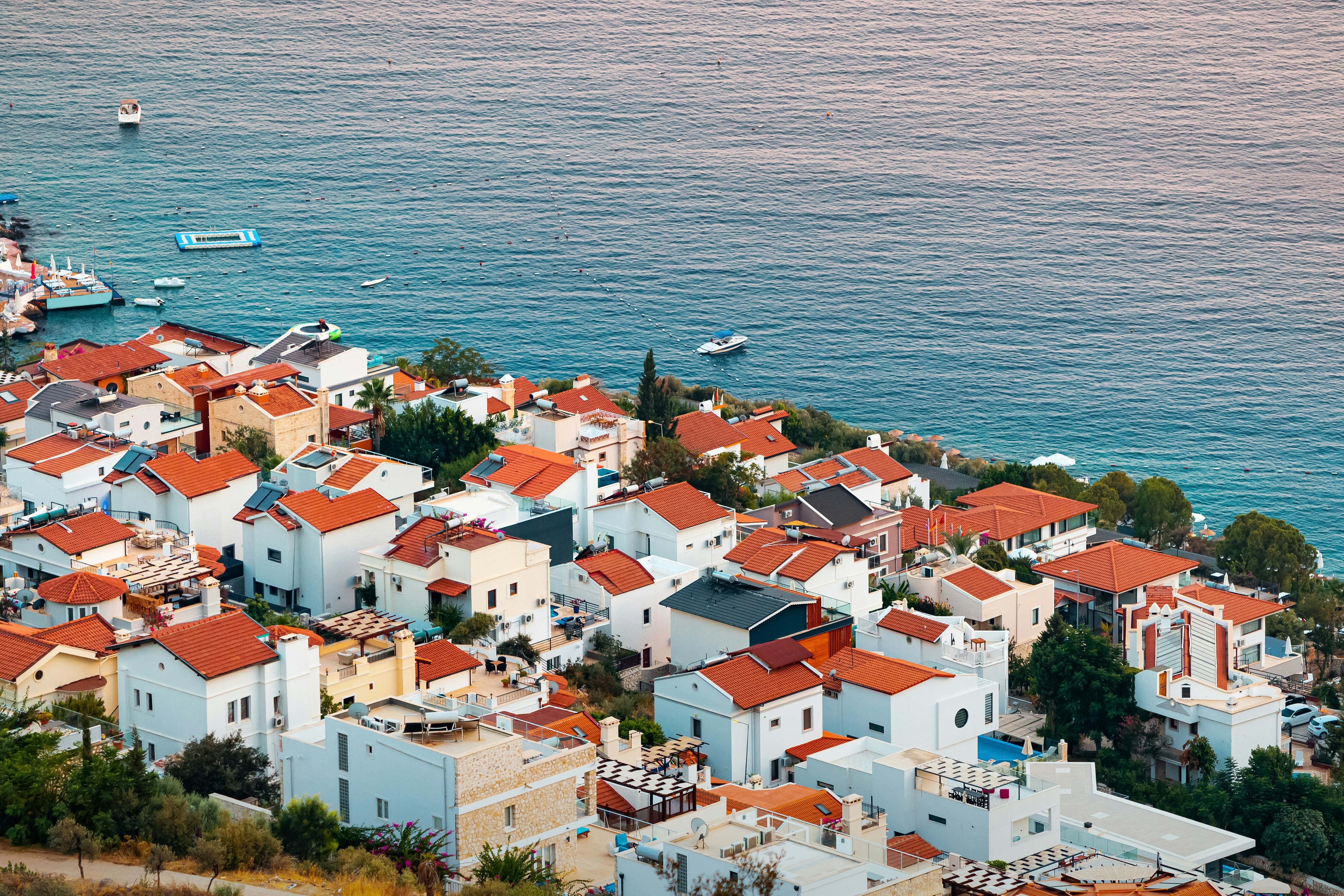 Aerial view of a Mediterranean coastal village with white buildings and red-tiled roofs by the sea, featuring boats and clear blue water.