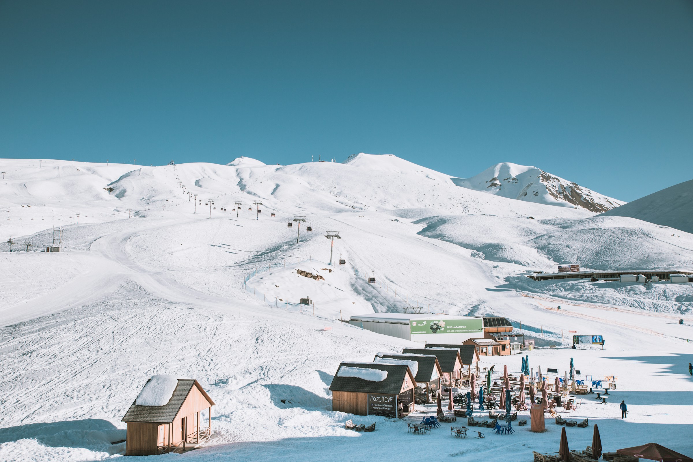Winter wonderland with snow-capped mountains and a ski resort with cabins and chairlifts under clear blue skies.