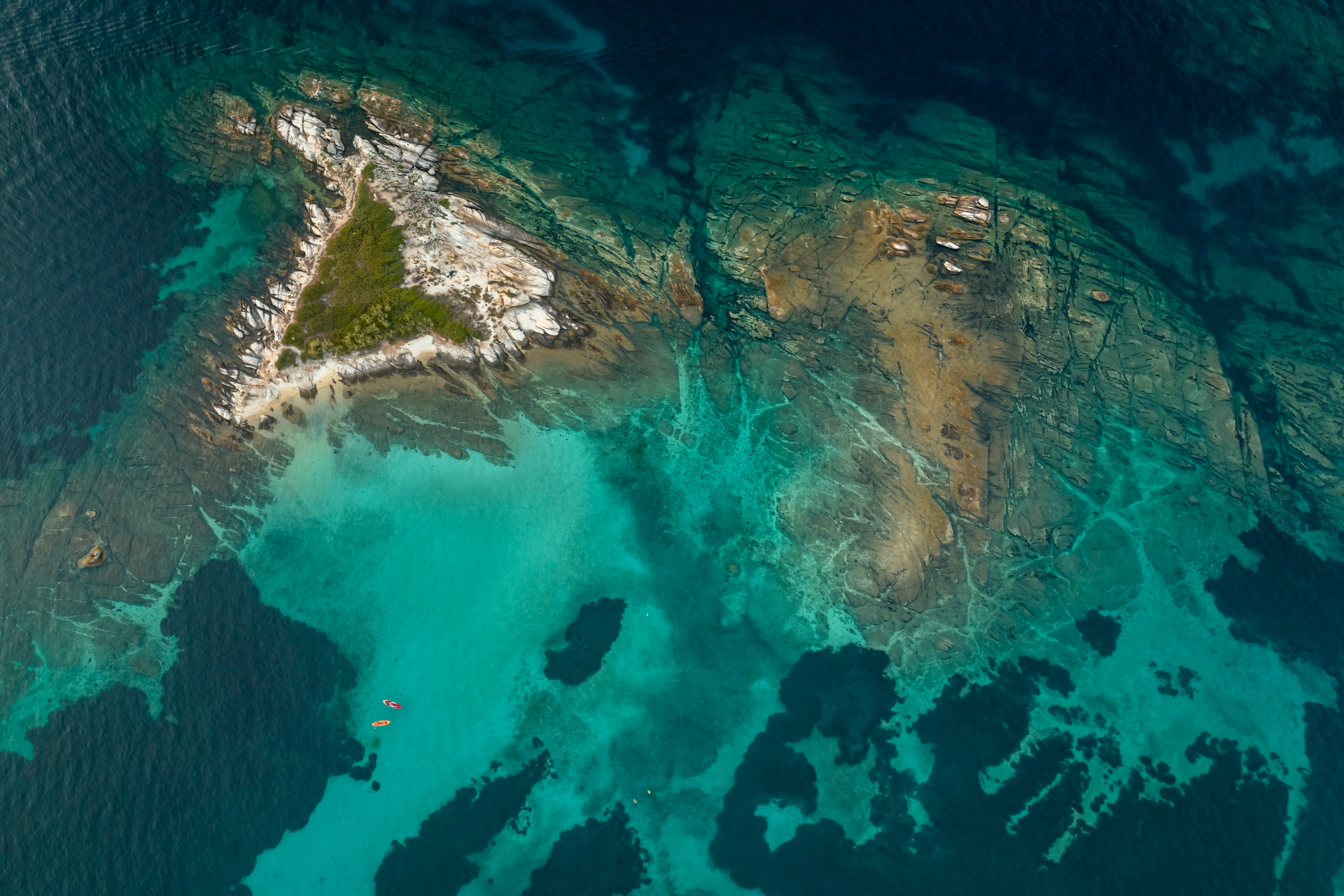 Aerial view of a triangular rocky island surrounded by vibrant turquoise sea, with patches of darker blue indicating deeper water.