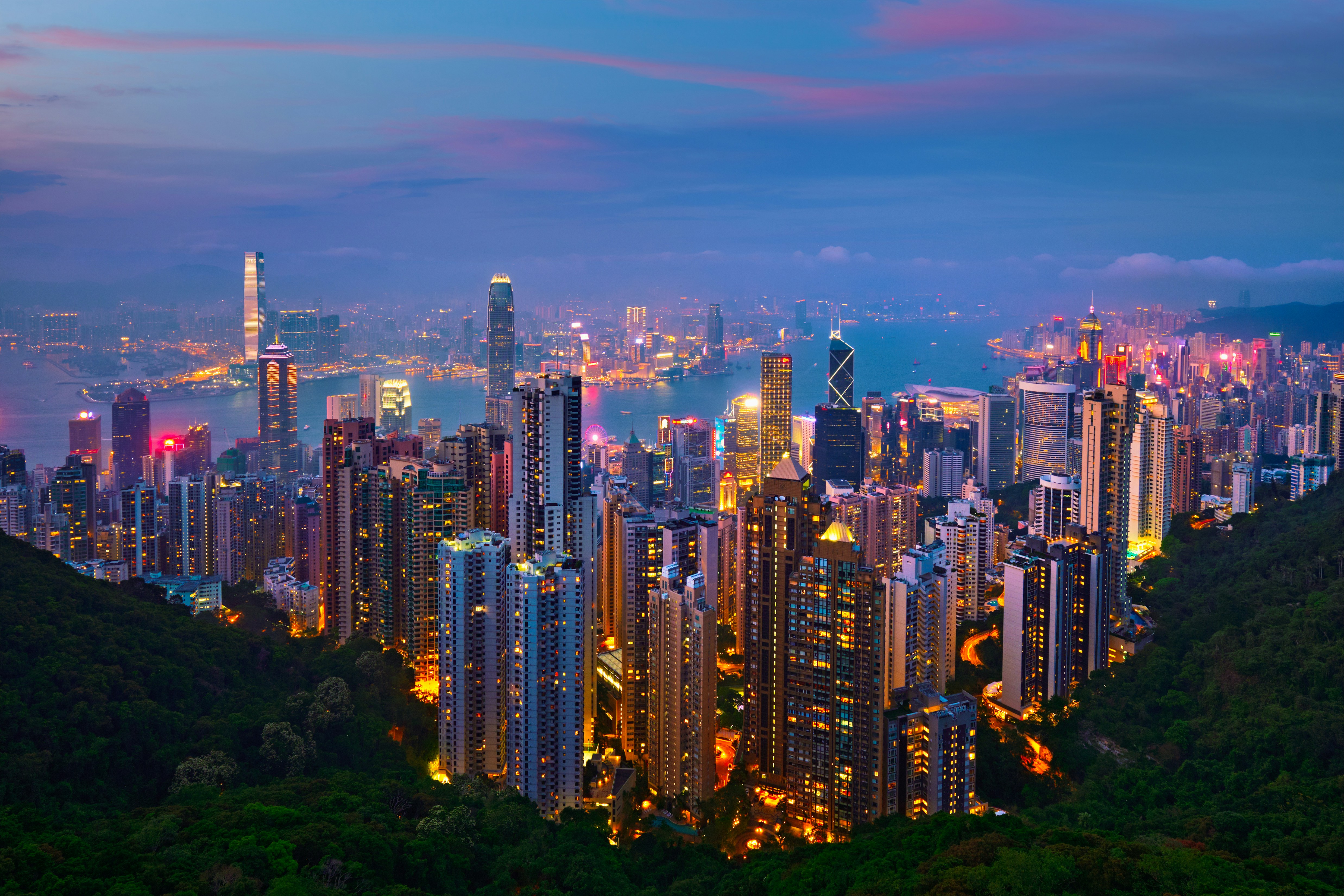 Hong Kong skyline at dusk with illuminated skyscrapers, Victoria Harbor, and surrounding hills, under a vibrant twilight sky.