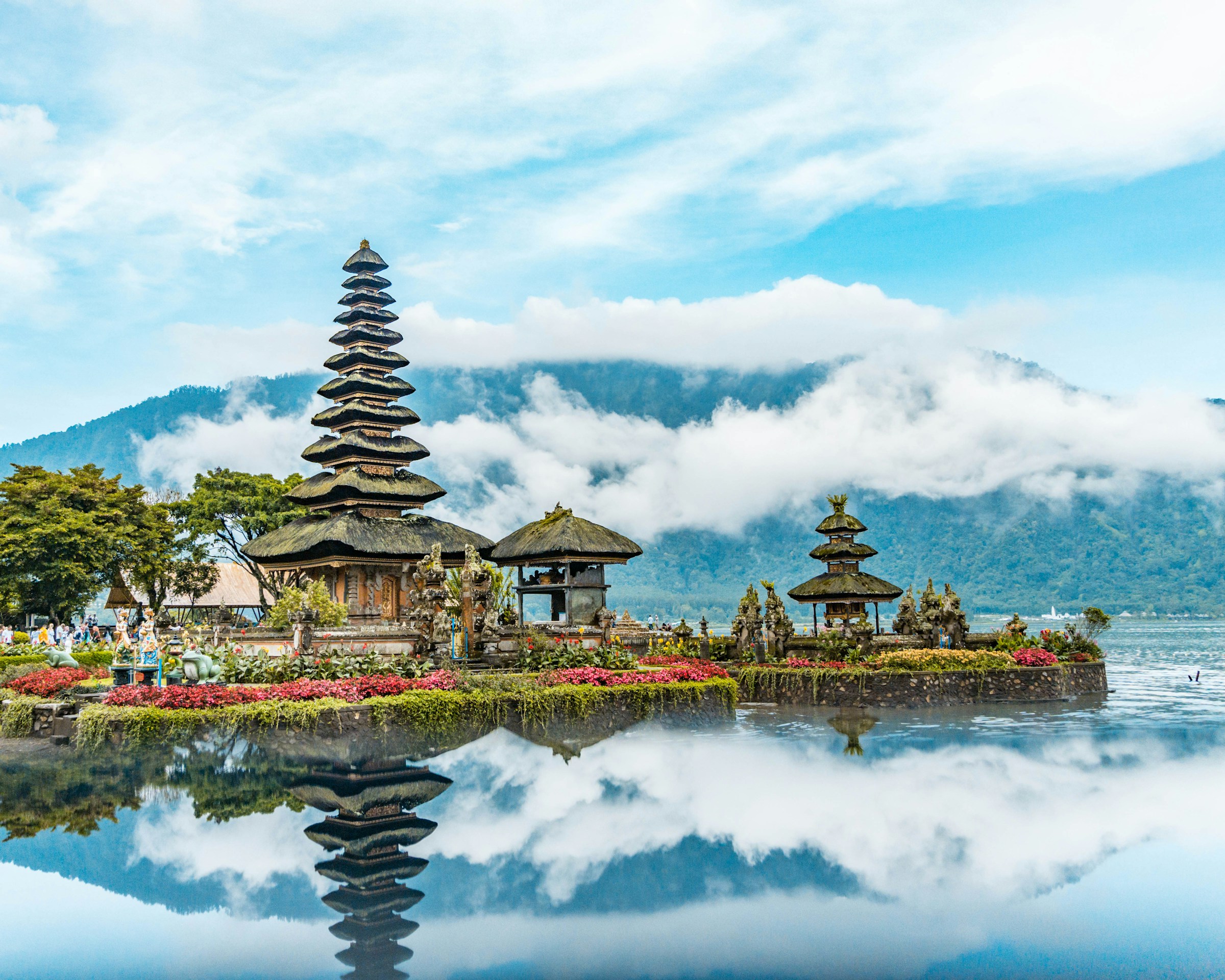 Balinese temple on Lake Bratan surrounded by lush greenery and mountains, with a clear blue sky and clouds reflected in the water.