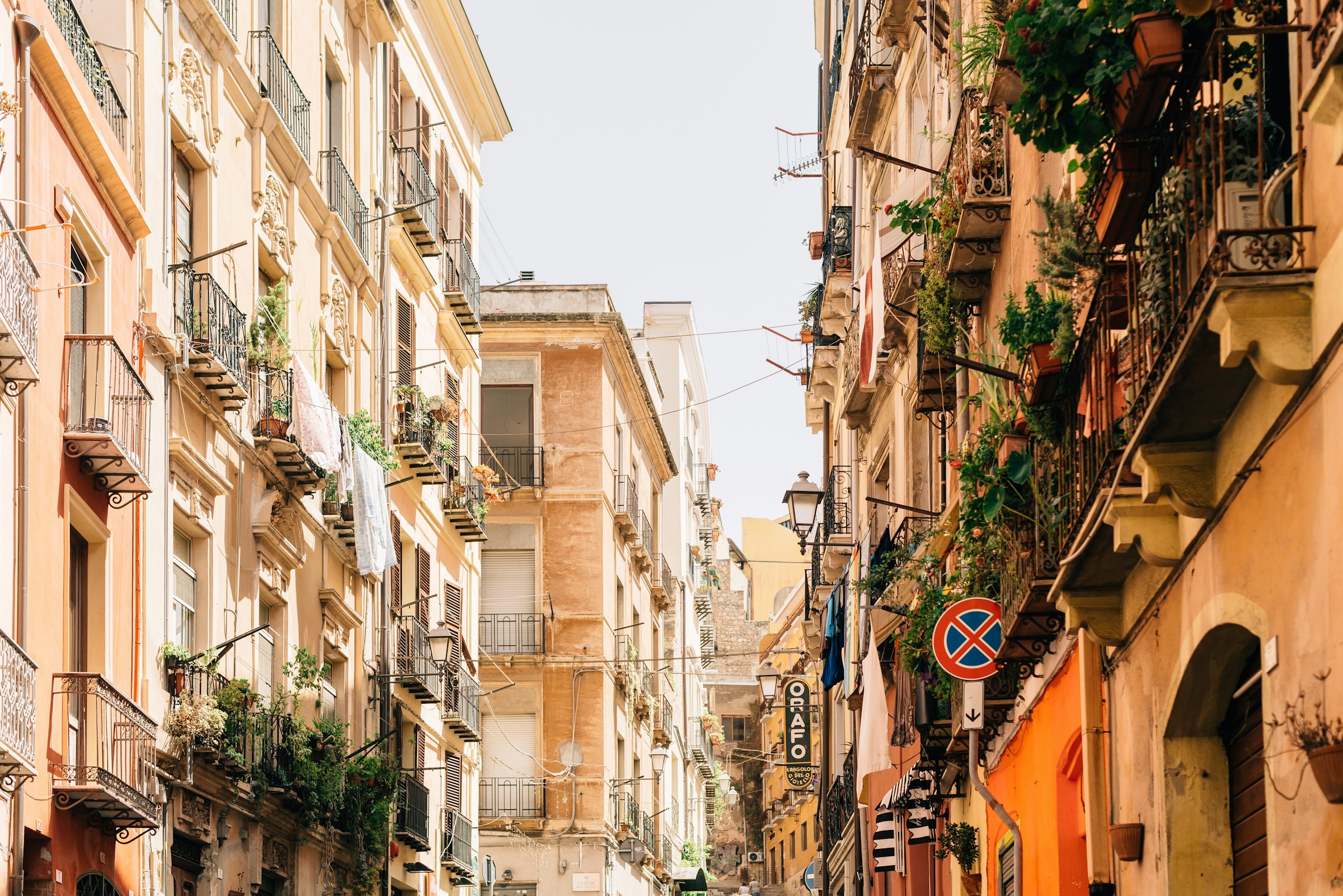 Narrow street in an Italian city lined with colorful old buildings adorned with balconies and plants, capturing a sunny day.