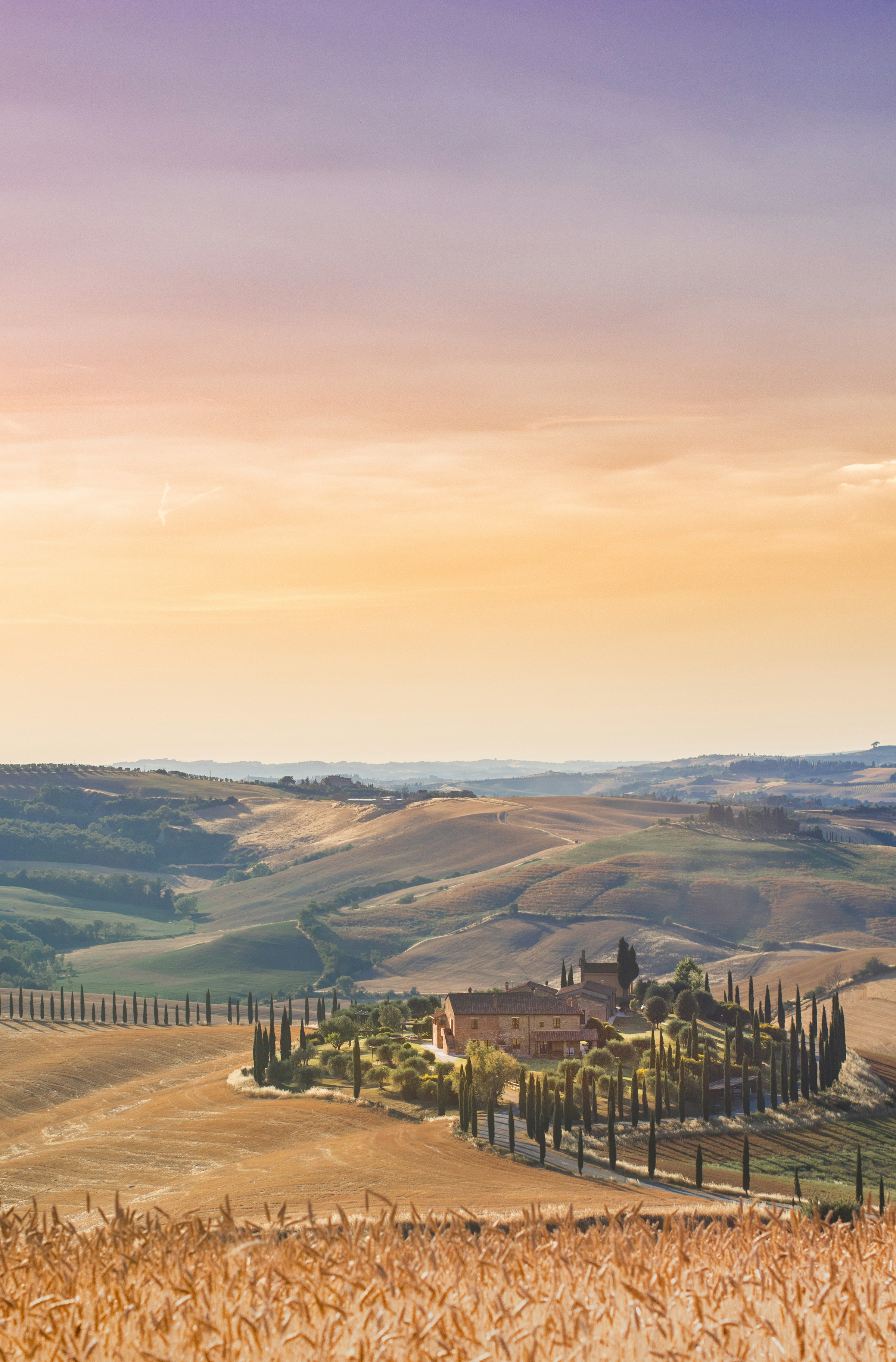 Sunset over rolling hills in Tuscany, Italy, with a traditional farmhouse surrounded by cypress trees and golden fields.