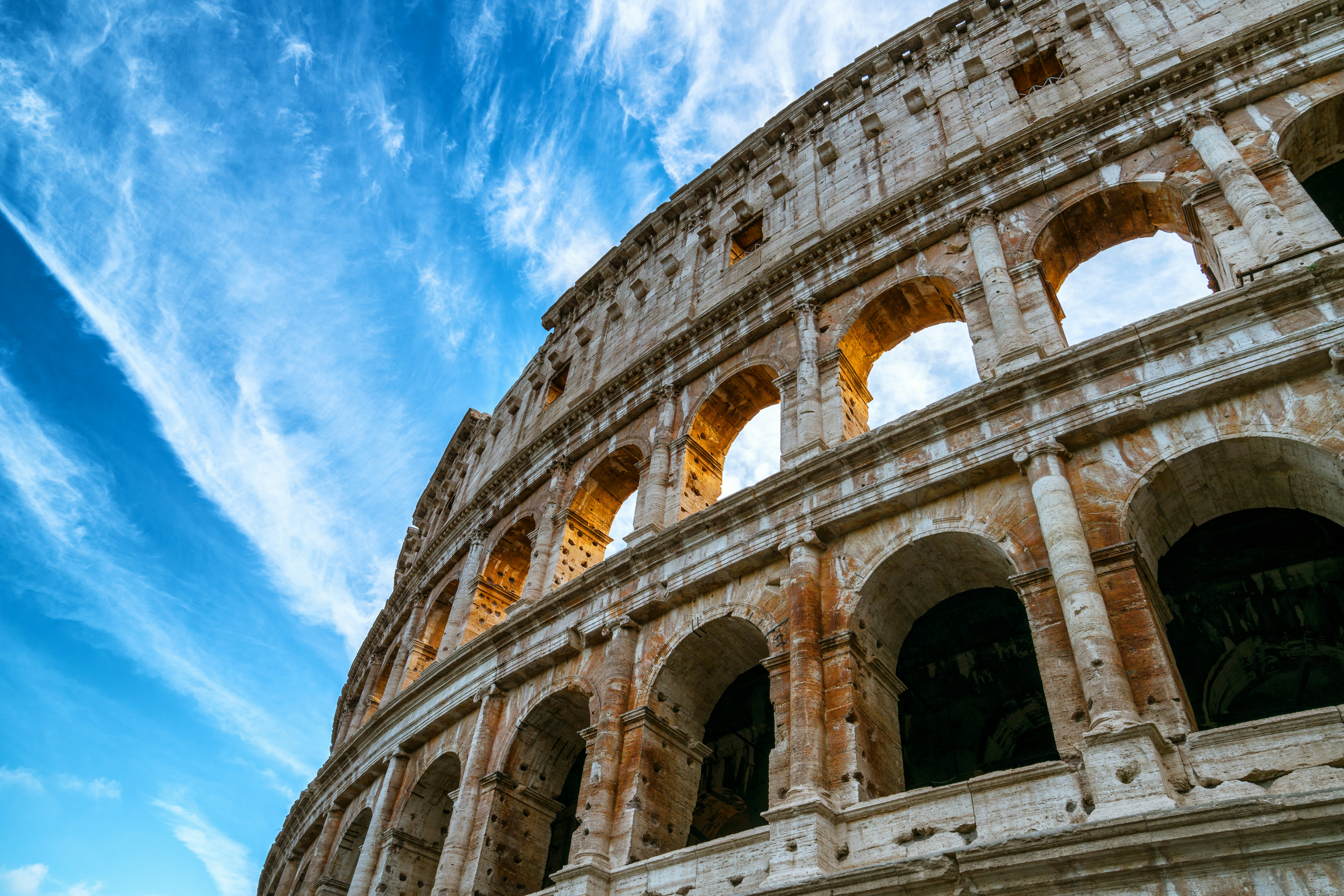 Exterior view of the Colosseum in Rome, showcasing its ancient arches and stone architecture against a bright blue sky.