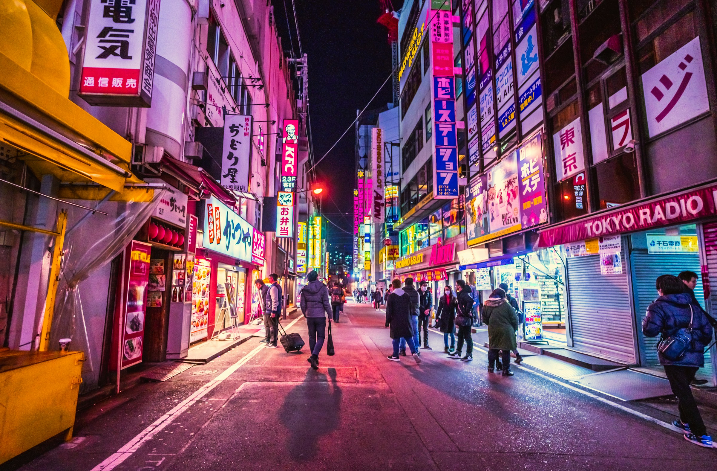 Street in Tokyo with neon signs lighting up all windows in pink light with people walking