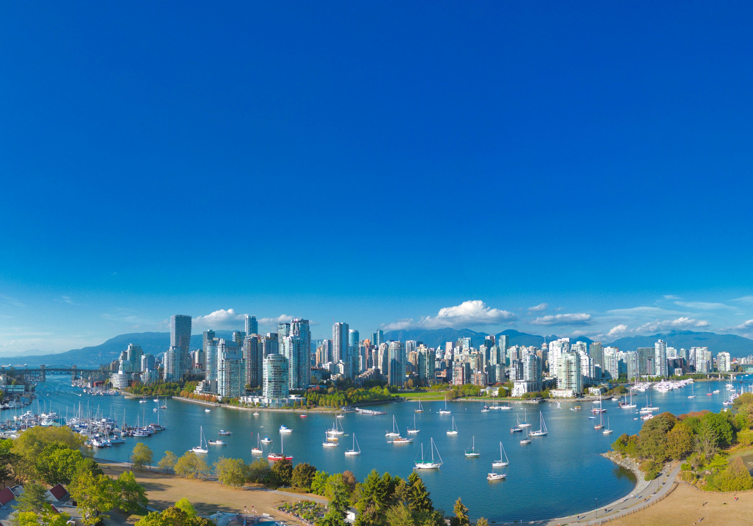 Vancouver skyline med skyskrapor vid vattnet och segelbåtar i hamnen under en klarblå himmel.