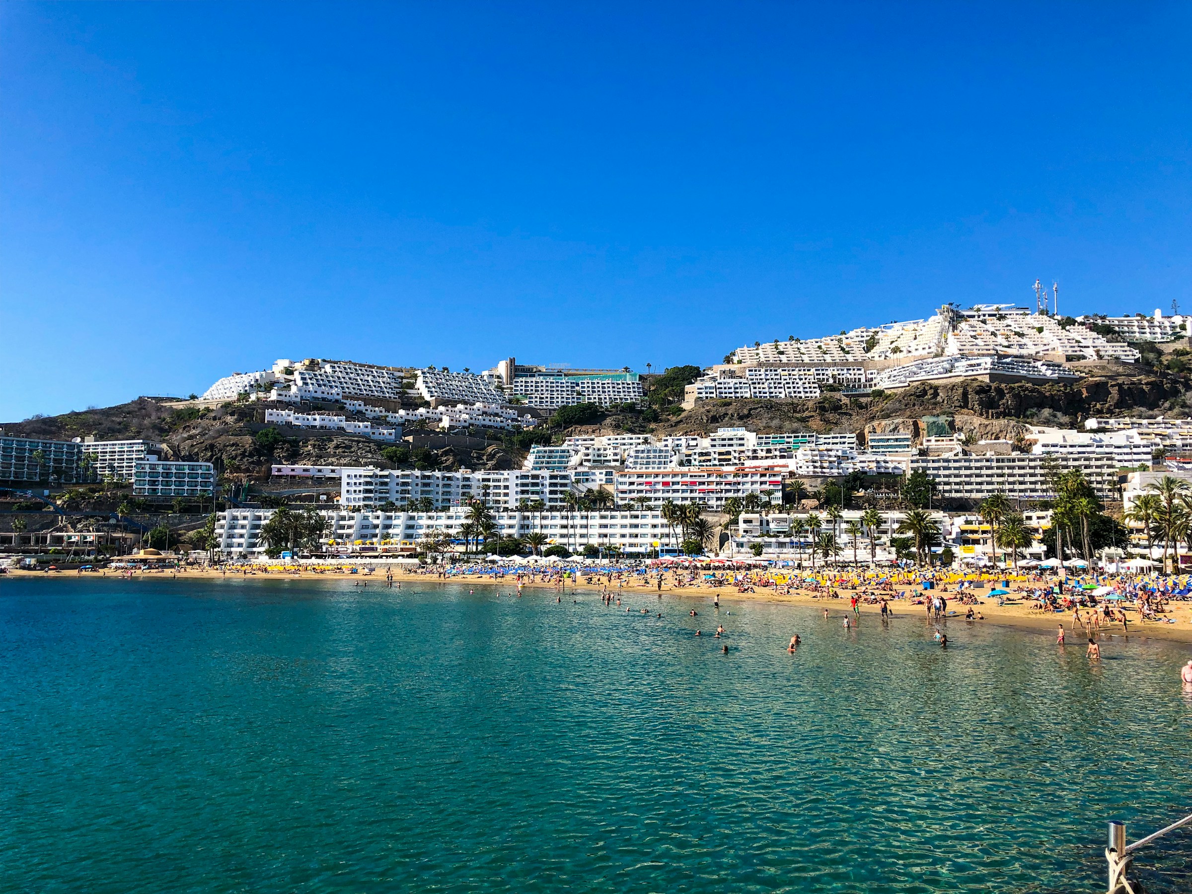 Beach and resort on the coast of Gran Canaria, Canary Islands, Spain, with turquoise sea, sandy beach and white high-rise buildings on a hill under clear blue skies.