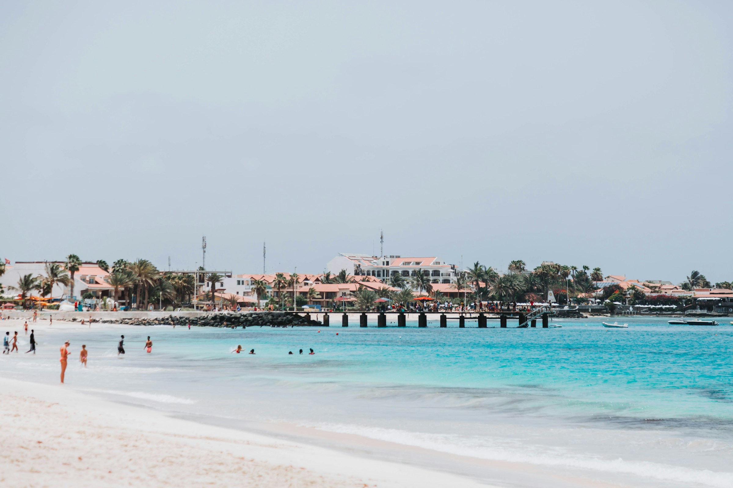 Travel to Sal - Aerial view of a wooden pier with people and red umbrellas overlooking turquoise ocean waters and sandy beach.